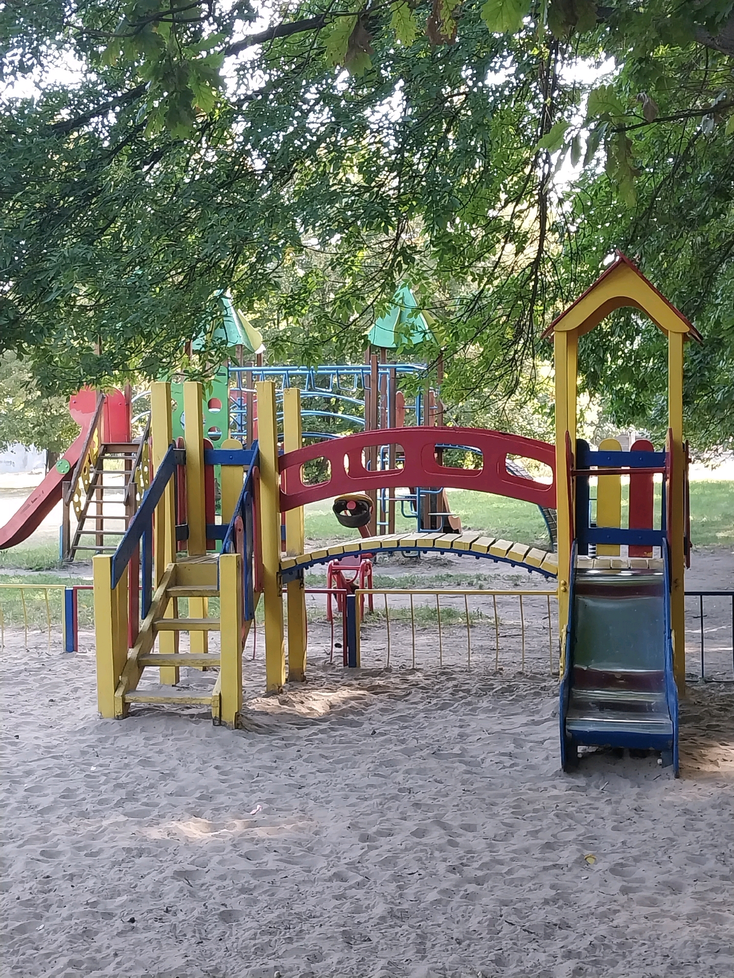 The image depicts a brightly colored children's playground located in Doslidne, Ukraine. The playground equipment is predominantly yellow and red, with accents of blue and green. Central to the scene is a multi-level structure featuring slides, stairs, and a bridge. To the left, a red slide descends from a platform. A wooden staircase with yellow steps and blue railings leads up to a higher section of the playground. A red archway, serving as a bridge, connects different parts of the structure. On the right, a blue and yellow tower houses a green slide. The playground is situated on a sandy surface, with a low yellow and blue metal fence surrounding parts of it. Behind the playground, green grass and trees are visible, with sunlight filtering through the leaves. The overall impression is of a well-maintained, outdoor recreational area for children.