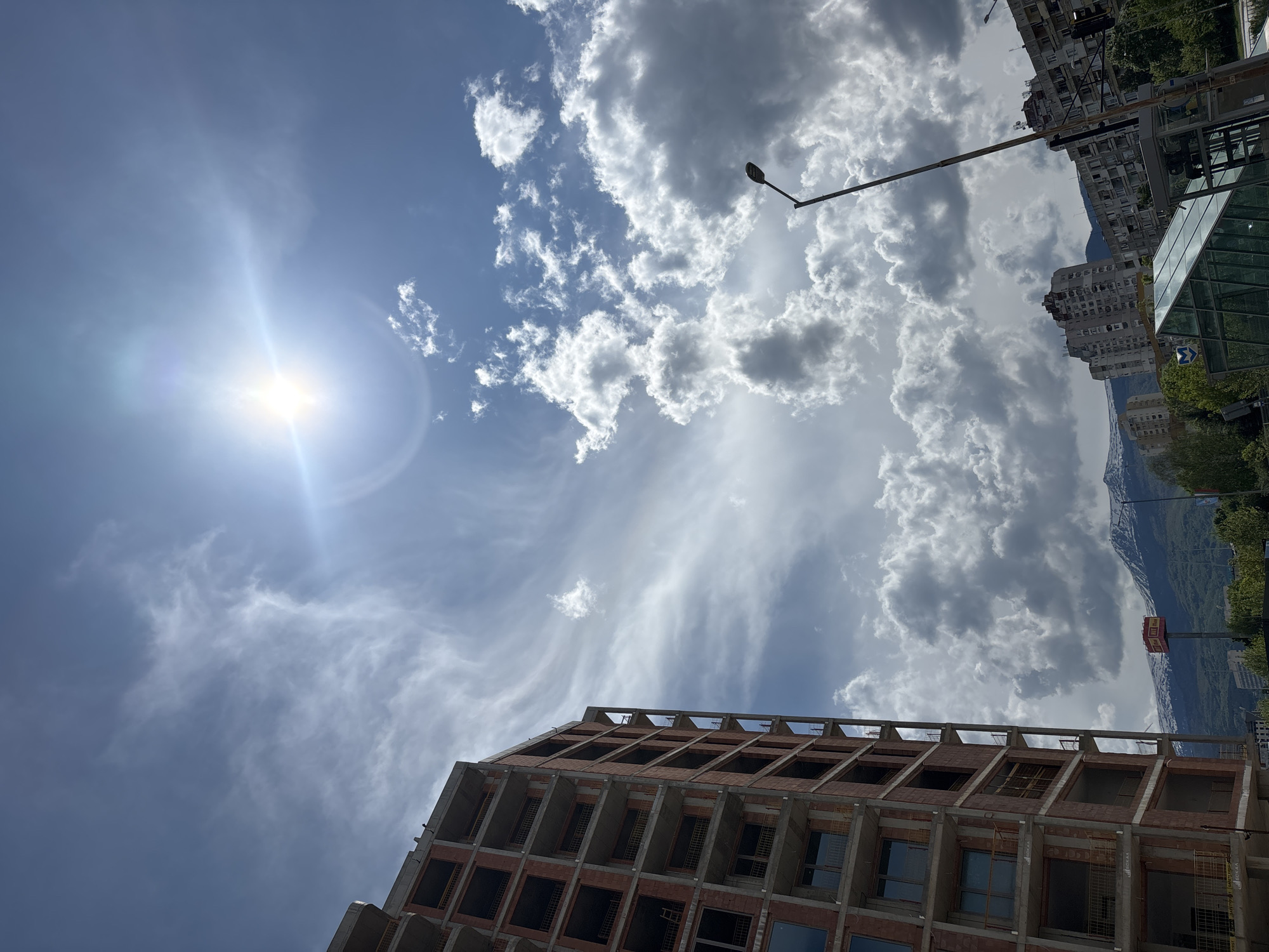 The image is taken from a low angle, looking up at a partly cloudy sky with the sun visible. The bright sun causes lens flare. In the foreground, the top of a building under construction is visible, featuring a red brick exterior and concrete framework. A street lamp is present, extending into the sky. In the background, other buildings are visible, as well as a mountain range. A sign is also visible in the background, though the text is unreadable.