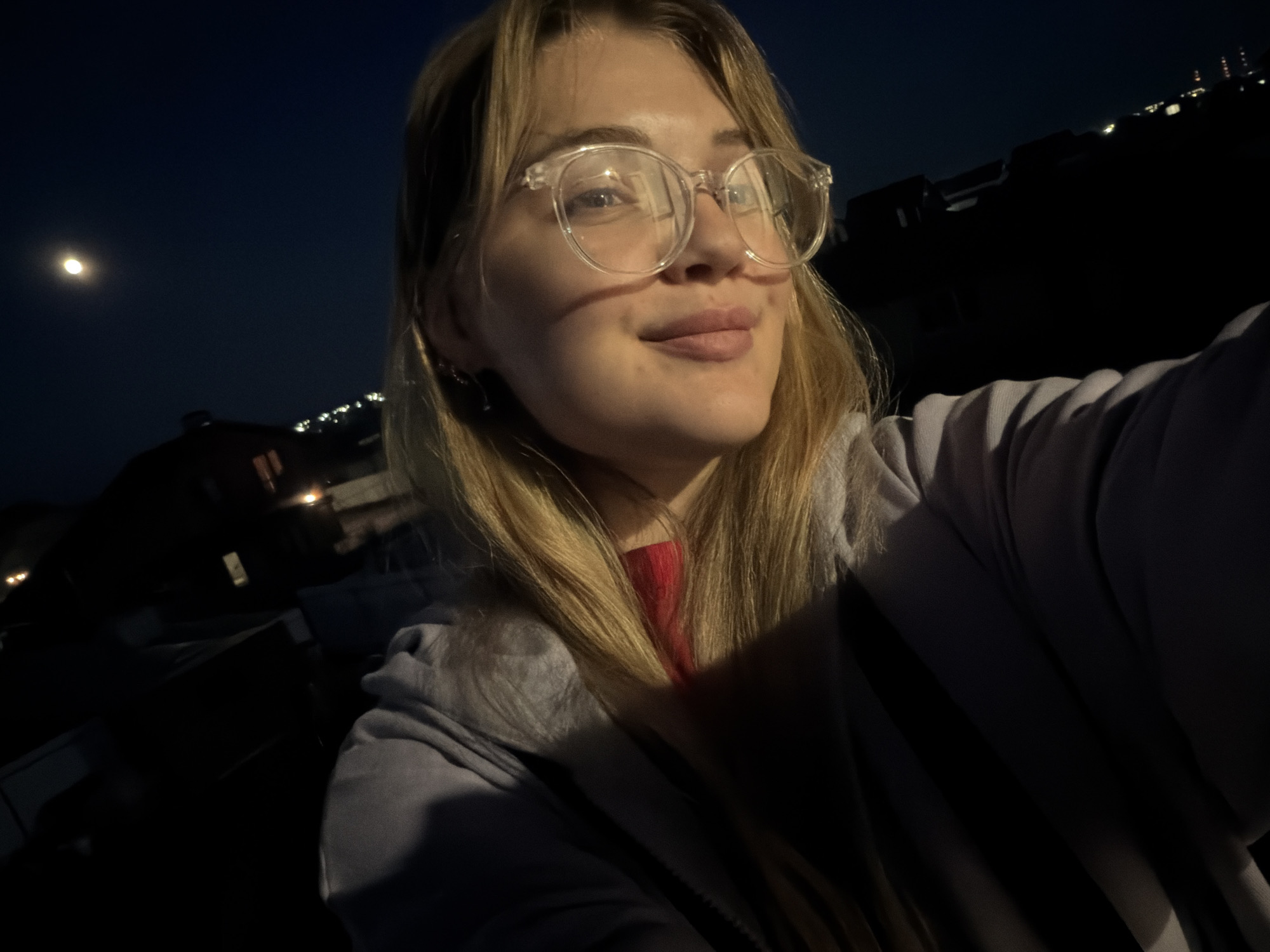 This close-up image is a night-time selfie featuring a young woman against a dark, expansive sky, likely taken in Большая Шильна, Russia. The woman, positioned in the foreground, has long, light brown hair and wears clear-rimmed glasses. She looks slightly upwards and to her right with a gentle, contented smile, her face and hair illuminated by an unseen light source. She is dressed in a light-colored hooded top, possibly grey or lavender, layered over a red garment.
The background clearly depicts a dark night sky with a prominent, bright moon visible in the upper left. Below the horizon, indistinct dark shapes suggest buildings or structures, with scattered faint lights indicating a distant settlement or industrial area. The scene conveys a sense of peacefulness and quiet, captured under a clear night sky. No text is visible in the image.
