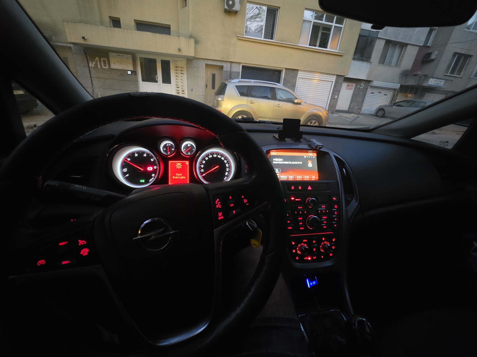 The image shows the interior of a car, likely an Opel, viewed from the driver's perspective. The dashboard is illuminated with red lighting. On the dashboard, a digital display shows the words "Check Brake Lights." Outside the car, a yellow SUV is parked in front of a residential building. The building appears to be an apartment complex with multiple windows and balconies. The image is taken during daylight, possibly late afternoon or early morning, given the lighting conditions. Based on the license plate style of the yellow SUV, and the architecture of the building, this scene is likely located in Varna, Bulgaria. The car is parked on a street, with other buildings and vehicles visible in the background. The overall scene depicts a common urban environment.