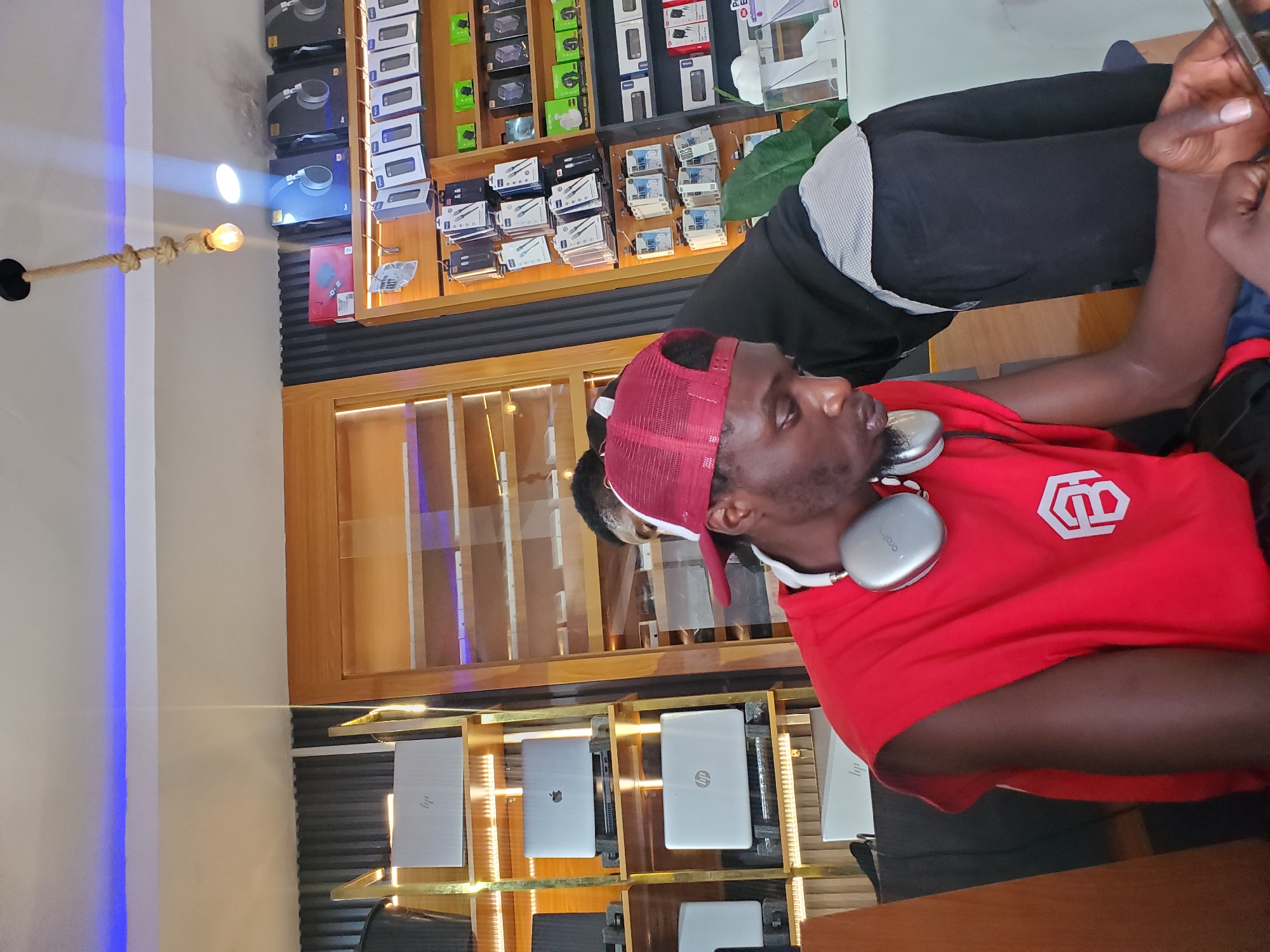 The image shows a young Black man wearing a red t-shirt and a red baseball cap, with headphones around his neck. He is seated in front of a display of laptops and other electronic devices, suggesting a retail or workshop environment. The man is looking towards the right of the frame, holding a smartphone in his right hand and appearing to be engaged with it.
The background is filled with shelves displaying various electronic items, including packaged headphones and what appear to be laptop boxes. The shelves are made of wood and have lighting that illuminates the products. There is also a section of what looks like a wall with a blue light strip and a rope light fixture with a bulb. The overall setting appears to be an electronics store or a similar commercial space. The lighting in the image is a mix of ambient store lighting and the light from the blue strip and rope light. The scene is well-lit, but the specific time of day is not discernible from the image. The man's posture and focus on his phone suggest a moment of personal engagement within the commercial setting. The context provided indicates the location is Jos, Nigeria.