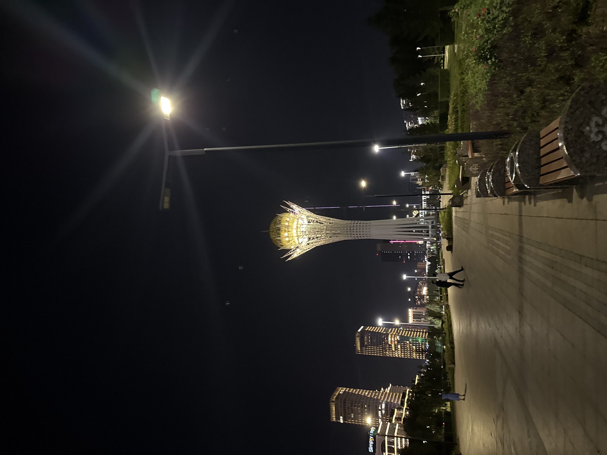 In the city of Astana, Kazakhstan, at night, a wide, paved walkway curves through a park. To the left, several modern, illuminated skyscrapers stand tall, one bearing the "Simply Implies" logo. Dominating the center of the frame is the Bayterek Tower, a distinctive landmark with a golden, sphere-like structure at its apex, illuminated and appearing to rise from a white, stylized stem. A tall, dark lamppost with a bright light stands in the foreground, casting a glow on the walkway. Several park benches with ornate stone bases and wooden slats are arranged along the path. To the right of the walkway, dense, manicured greenery and flowering plants form a park area. Two individuals are walking away from the camera on the walkway, dressed in dark clothing. Another person, wearing a light blue dress, stands alone further down the path. The sky is dark, with light flares emanating from the lamppost.