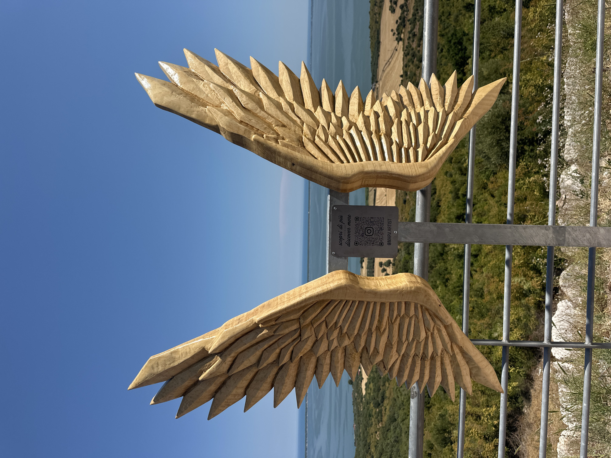 An outdoor shot shows a pair of large, carved wooden wings, displayed vertically against a clear blue sky and a scenic background of water and vegetation. The wings are intricately detailed, with each feather meticulously carved to create a textured, realistic effect. They appear to be mounted on a metal pole, which is also part of a metal railing in the foreground.
Attached to the support structure for the wings is a small metal plaque. The plaque contains text in Italian: "scopri di più discover more". Below this text, a QR code is visible, along with an Instagram logo and the handle "@MARV.ARTIST".
The background features a body of water, likely a lagoon or bay, stretching towards the horizon. On the far shore, there is a landscape of dry, rolling hills dotted with greenery and some cultivated fields. The overall scene suggests a scenic viewpoint or an art installation in a natural setting.
The location is identified as Cagnano Varano, Italy, a town known for its proximity to the Gargano National Park and the Lago di Varano. This setting is consistent with the visual elements of the image. No people are present in the frame. The lighting indicates a bright, sunny day.