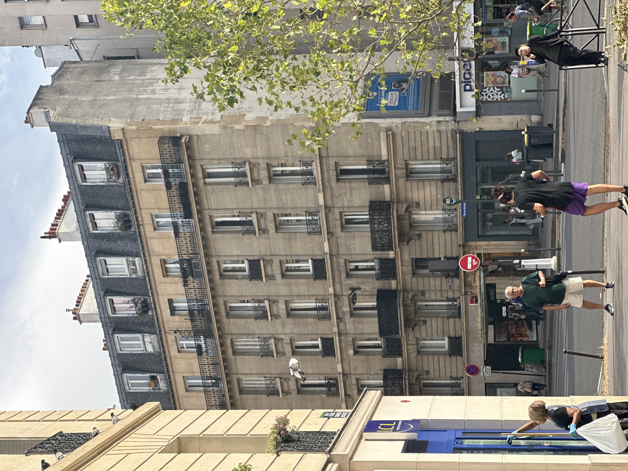 In Paris, France, a street scene unfolds under a cloudy sky. On the left, a cream-colored building with a black wrought-iron balcony displays potted pink flowers. To its right, a taller, multi-story building with a mansard roof and dormer windows dominates the frame. The building's facade is a neutral beige, punctuated by numerous windows with black wrought-iron railings on several floors.
In the foreground, a woman wearing a dark apron and blue gloves is cleaning a blue-tinted glass storefront, holding a white plastic bag. Next to her, a man with graying hair and wearing a green t-shirt and white shorts stands facing forward, carrying a black bag over his shoulder. Further down the street, a young man in a black t-shirt and purple shorts stands facing away from the camera, and another man in a black suit is on his phone. Various businesses and street signs are visible on the buildings lining the street. A green trash bin is situated near a bus stop sign. A mature tree with green leaves partially obscures the right side of the buildings. The street is paved, with a sidewalk in the foreground.