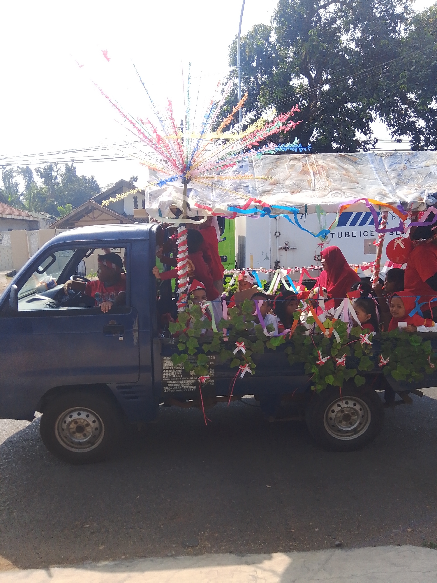 In Kalinyamatan, Indonesia, a flatbed truck is decorated for a parade. The truck bed is laden with children and adults, many wearing red clothing. The passengers are adorned with festive decorations, including colorful ribbons and garlands of greenery. A large, ornate centerpiece, resembling a tree or a burst of fireworks made of colorful streamers, is erected in the middle of the truck bed. The truck itself is blue, and the driver, wearing a red shirt, is visible behind the steering wheel. The truck is decorated with a white tarp that covers the top and sides, adorned with more streamers. In the background, there are houses and trees, suggesting a residential area. The truck appears to be in motion, participating in a celebratory event.
