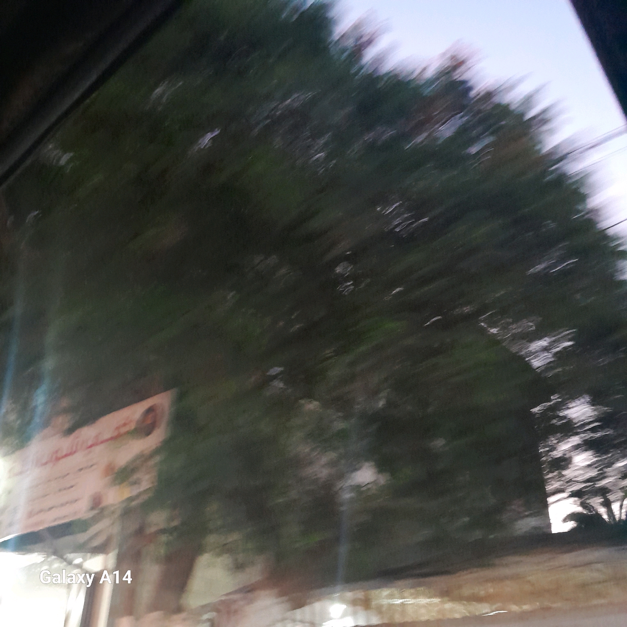The image depicts a blurry scene, likely taken from inside a moving vehicle, showing a tree with dark green foliage against a pale sky. A horizontal sign, partially visible and also blurred, is positioned in the lower left quadrant of the image. Text is present on the sign, with some Arabic script visible. The bottom left corner of the image contains the text "Galaxy A14". The overall impression is that of a street view captured during motion in the city of Deir el-Balah, Palestinian Territories.
