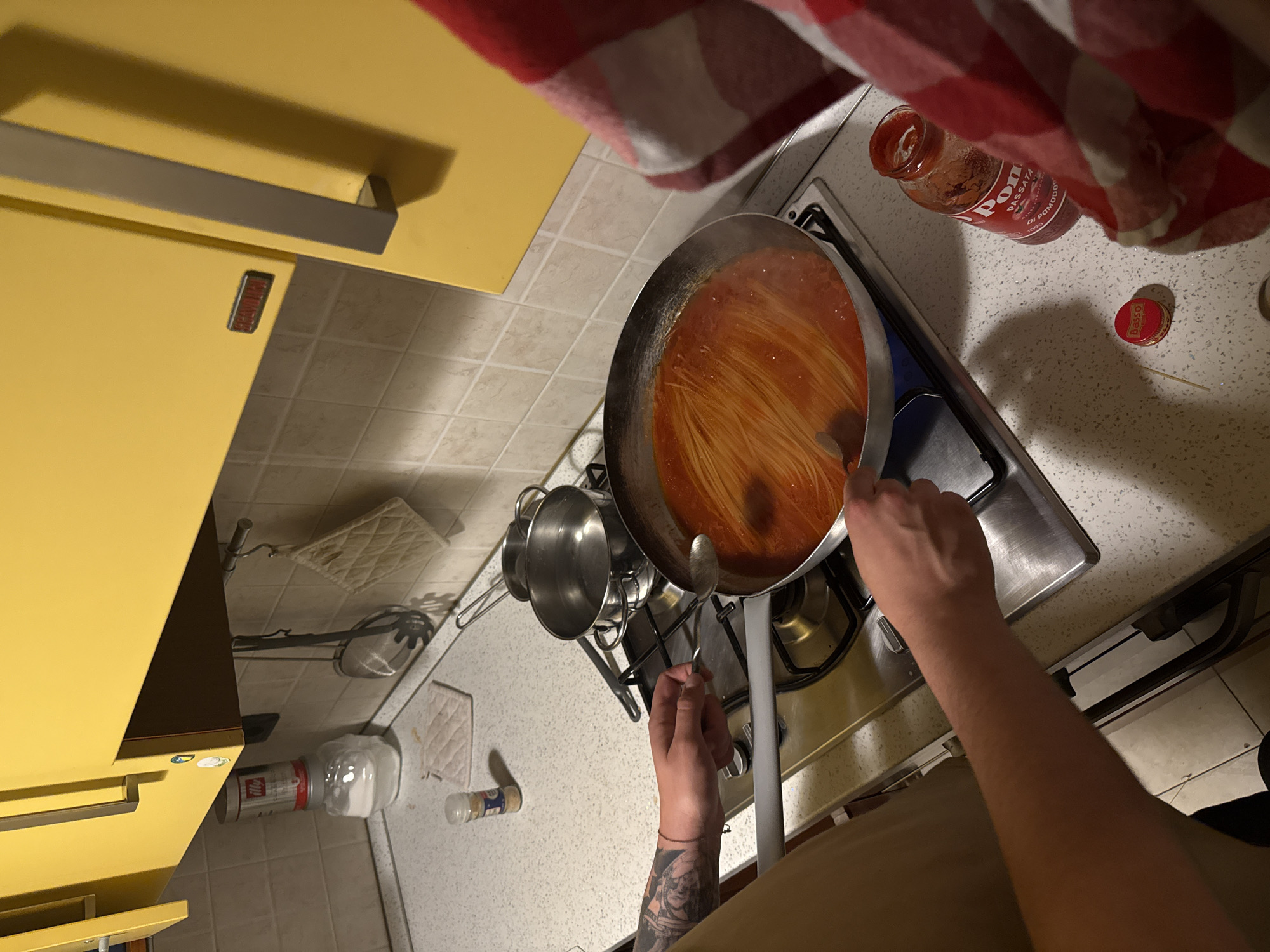 A person with a tattoo on their left forearm is cooking spaghetti with tomato sauce in a silver pan on a gas stove. The person is holding a silver spoon in their right hand. Next to the stove, there is a jar of tomato sauce and a red and white checkered cloth. On the counter to the left, there are various containers, including a white container, and a yellow kitchen cabinet above. The image was most likely taken in Milan, Italy, due to the "Dasso" brand on the tomato sauce jar, which is an Italian brand.