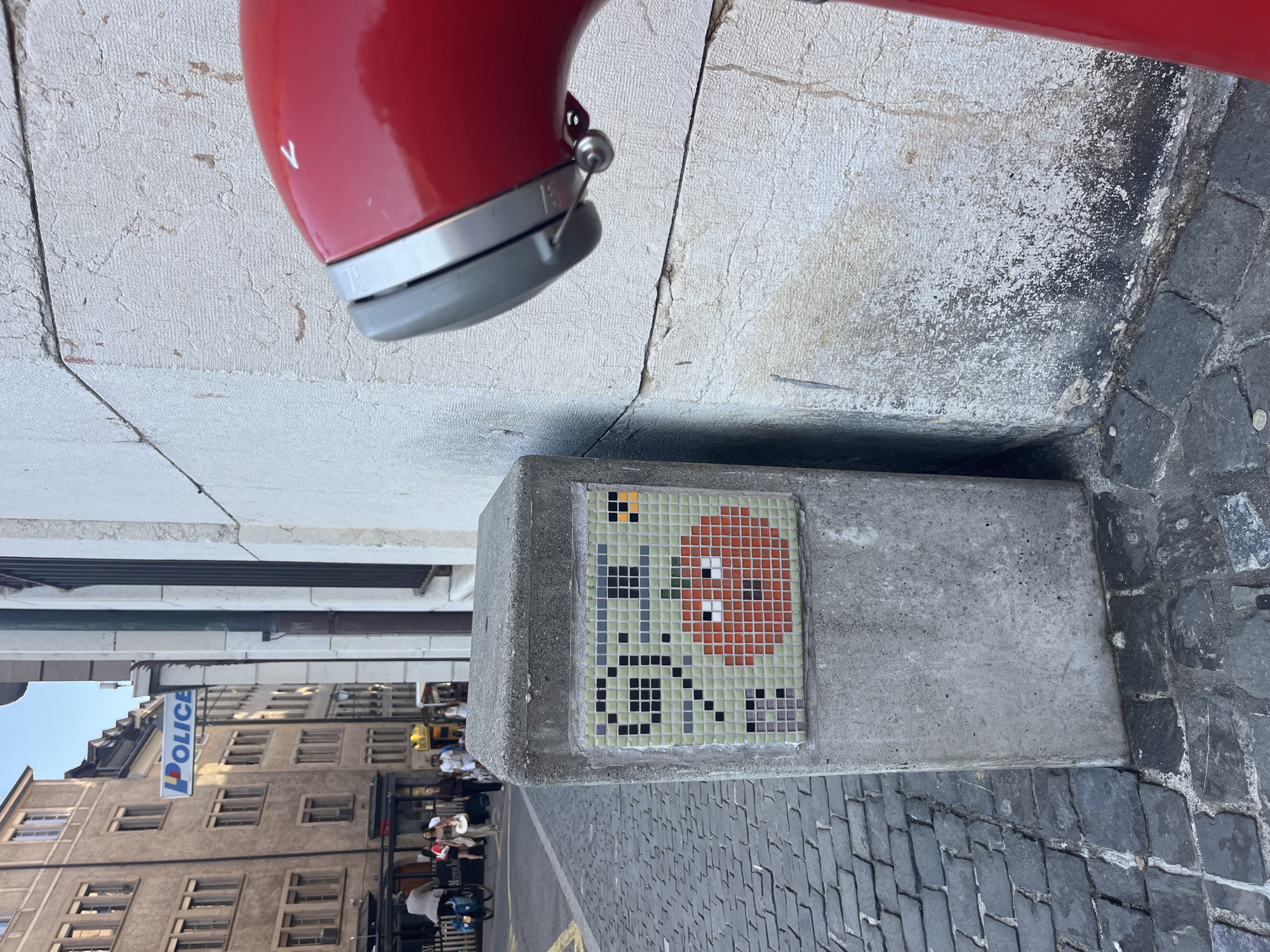 A low-angle, close-up shot captures a section of a cobblestone street in Geneva, Switzerland, with a concrete pillar in the foreground adorned with a pixelated mosaic of a character. The mosaic depicts a green figure with black eyes and a wide, red smile, reminiscent of a classic video game character. To the left of the pillar, a sign for "POLICE" is visible, mounted on a building. The street is lined with traditional European architecture, and pedestrians are seen in the background. A bright red object, possibly a fire hydrant or part of a public utility, occupies the right side of the frame, partially obscuring the building wall. The scene is illuminated by natural daylight, casting shadows and highlighting the textures of the stone pavement and building facades.