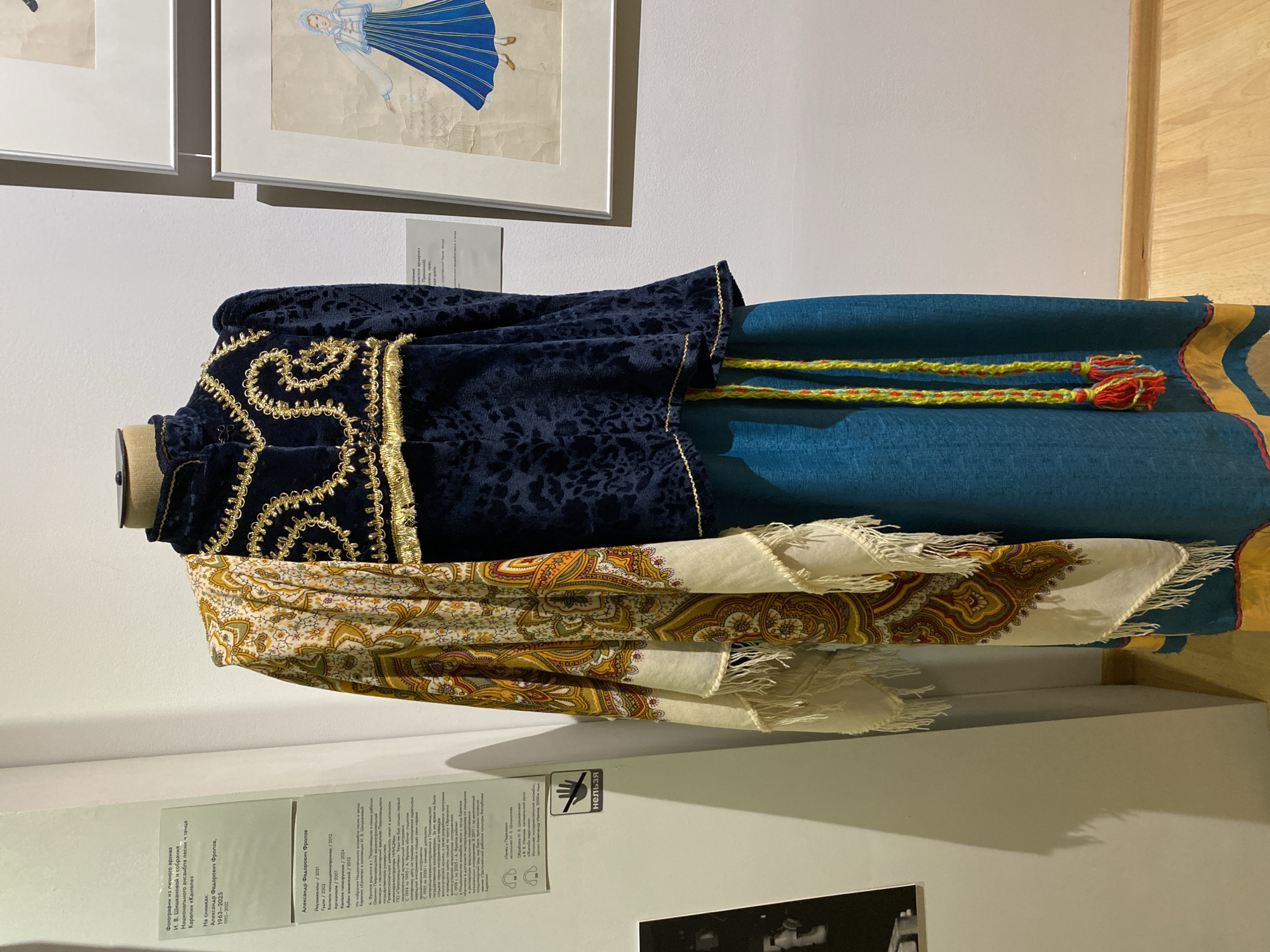 The image shows a mannequin displaying a traditional costume in what appears to be a museum exhibition. The costume consists of a dark blue velvet jacket with intricate gold embroidery on the collar and chest, a patterned shawl draped over the shoulders, and a long teal skirt with a yellow and blue woven belt. The mannequin stands in front of a white wall with several framed artworks displayed to the right. To the left, there is a display panel with text in Russian and a "НЕЛЬЗЯ" (forbidden) sign with a hand icon. The overall environment suggests an indoor exhibition space. The text on the display panel mentions Alexander Fyodorovich Frolov and provides dates, implying the exhibition is dedicated to his work or collection. Research indicates that Alexander Frolov was a significant figure in the preservation of Karelian culture and traditions, and his archives are housed in the National Museum of the Republic of Karelia, located in Petrozavodsk, Russia. Therefore, this image is likely from an exhibition at the National Museum of the Republic of Karelia in Petrozavodsk.