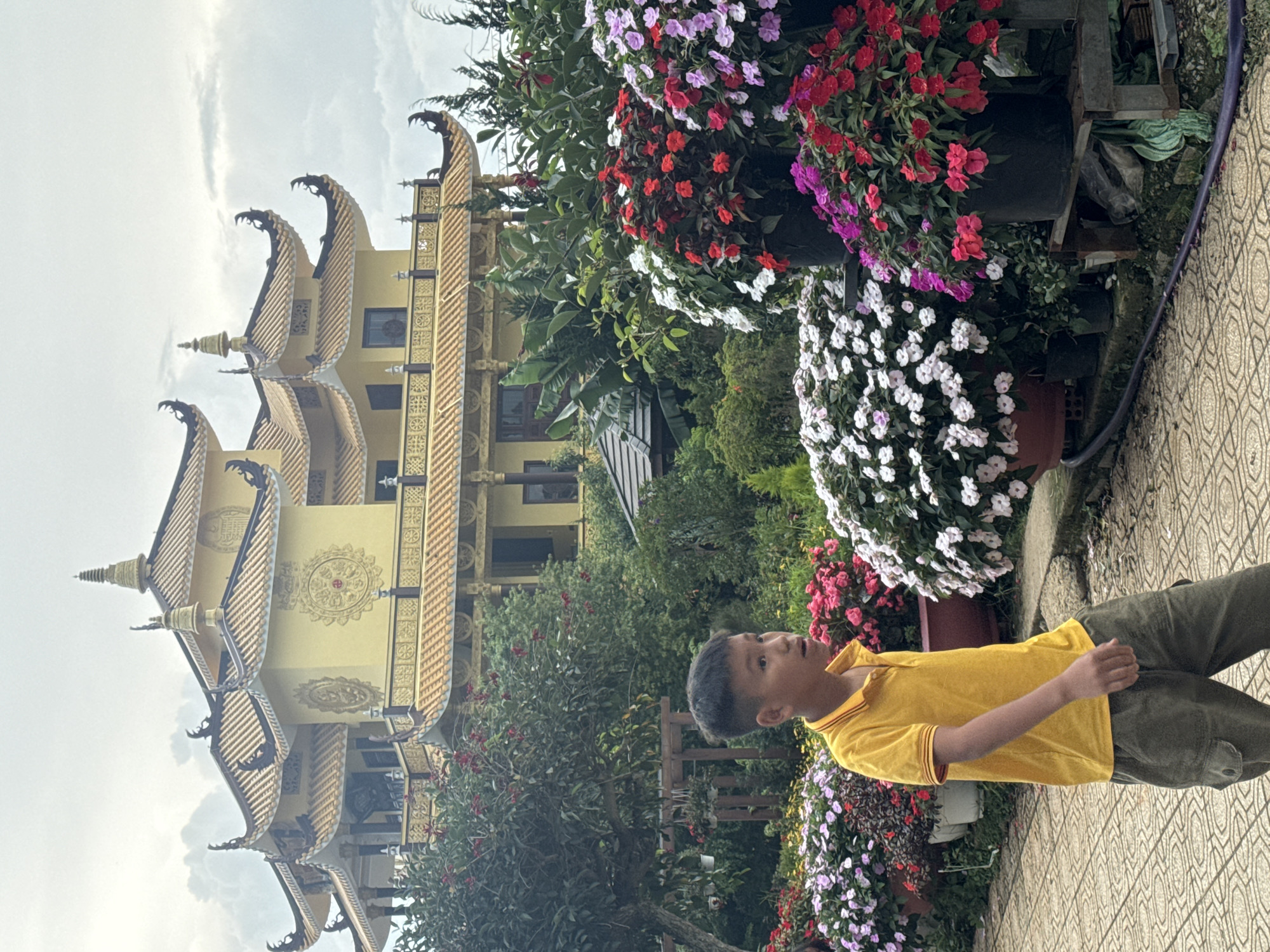 The image shows a young boy in a yellow polo shirt and green cargo pants walking on a patterned tiled path. He is in front of a vibrant display of flowering plants in various colors, including pink, red, and white. In the background, a large, ornate yellow building with multiple tiers and curved, tiled roofs stands prominently. This building is characteristic of Vietnamese Buddhist temples, and the lush floral arrangements suggest a garden setting. The sky is overcast. The overall scene is likely taken in Da Lat, Vietnam, a city known for its botanical gardens, temples, and French colonial architecture. Specifically, the architectural style of the building strongly resembles the Linh Phuoc Pagoda in Da Lat, a famous landmark renowned for its intricate mosaic work and distinctive tiered roof.