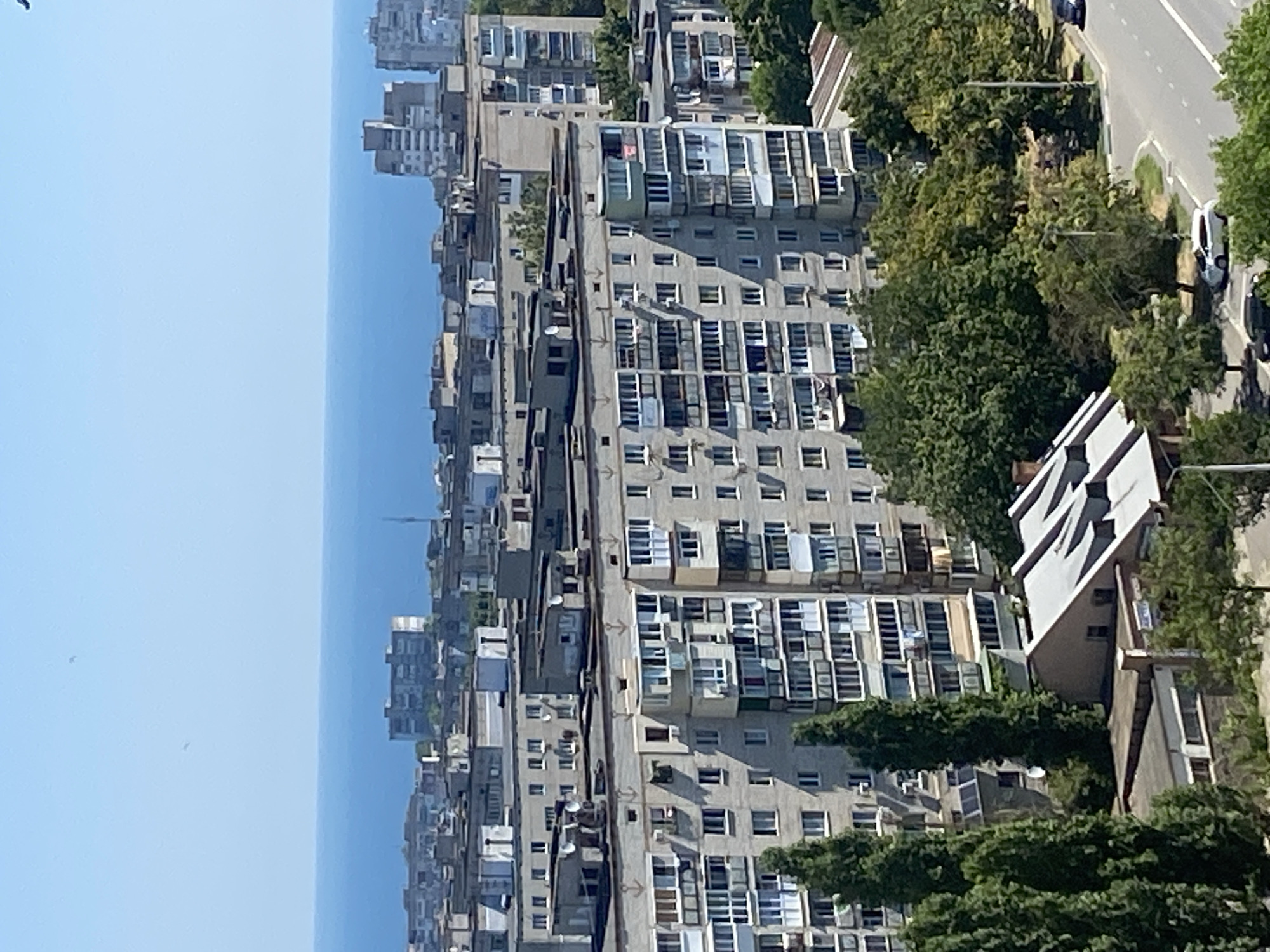 An aerial view captures a section of Chornomorsk, Ukraine, on a clear, sunny day. The horizon is dominated by the expansive, calm blue sea, meeting a pale blue sky. In the foreground, a cluster of Soviet-era apartment buildings stretches across the frame, characterized by their uniform grey facades, numerous balconies, and multi-story design. Some balconies are enclosed, while others are open, with laundry visible on some. Green trees, some tall and slender, others with fuller canopies, are interspersed between the buildings and in the lower sections of the image, adding a touch of nature to the urban landscape. A street is visible in the bottom right, with a few cars parked and a single white car driving on it. The architecture suggests a coastal city setting with a strong Soviet influence.