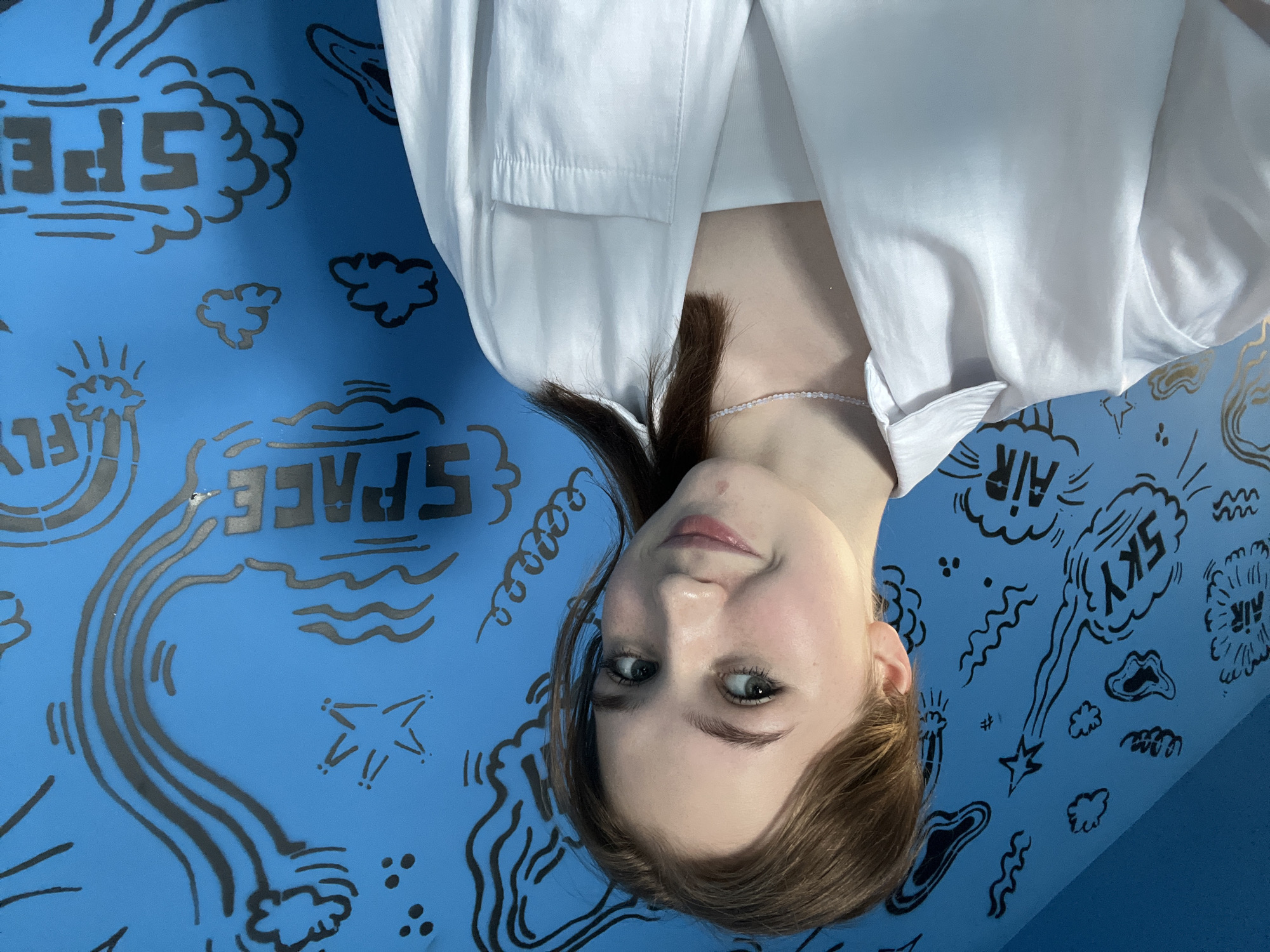 A young woman with fair skin and brown hair is positioned in front of a blue wall decorated with black drawings and words related to space and flight. She is wearing a white shirt. The background includes words like "SPACE," "FLY," "SKY," and "AIR" rendered in a stencil style, alongside illustrations of clouds, stars, and stylized lines suggestive of movement or trajectories. The woman's face is slightly tilted. The location is Izhevsk, Russia.