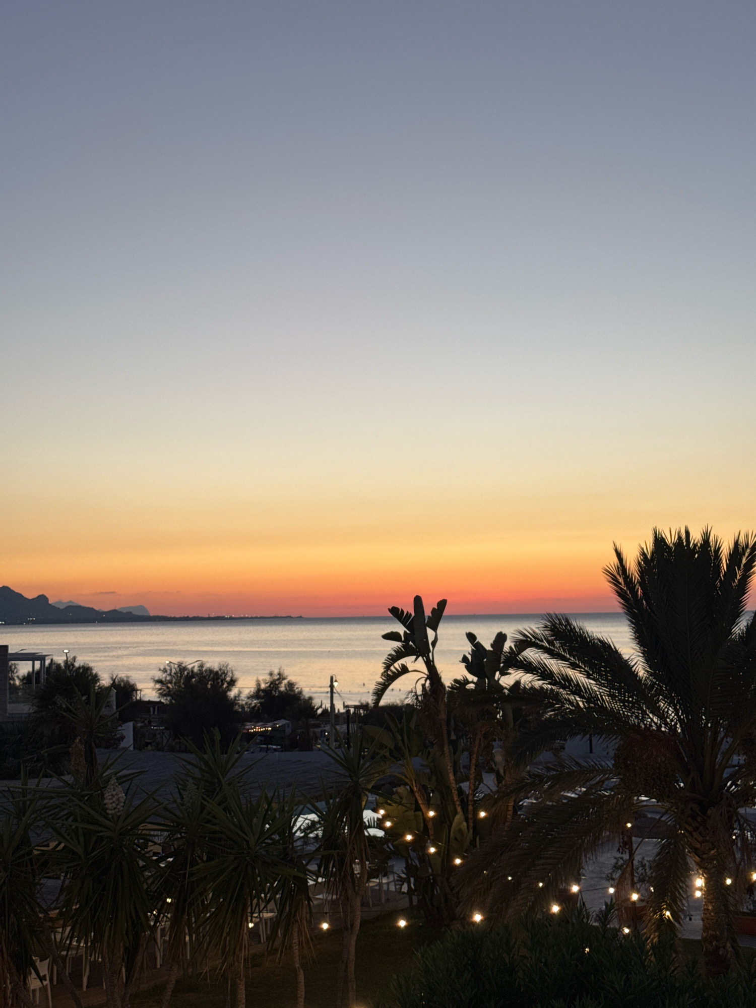 The image captures a serene twilight scene at a beachside location in Isola delle Femmine, Italy. In the foreground, silhouetted palm trees and other lush vegetation are illuminated by small, scattered string lights, creating a warm, inviting ambiance. Beyond the foliage, a calm sea stretches towards a distant coastline dotted with buildings and framed by undulating hills or mountains. The sky transitions from a soft pale blue at the top to a vibrant gradient of orange and red near the horizon, indicative of a sunset or sunrise. A thin strip of sand separates the sea from the vegetated area, where tables and chairs are visible, suggesting a restaurant or outdoor seating area. The overall impression is one of tranquility and natural beauty, characteristic of a Mediterranean coastal resort.