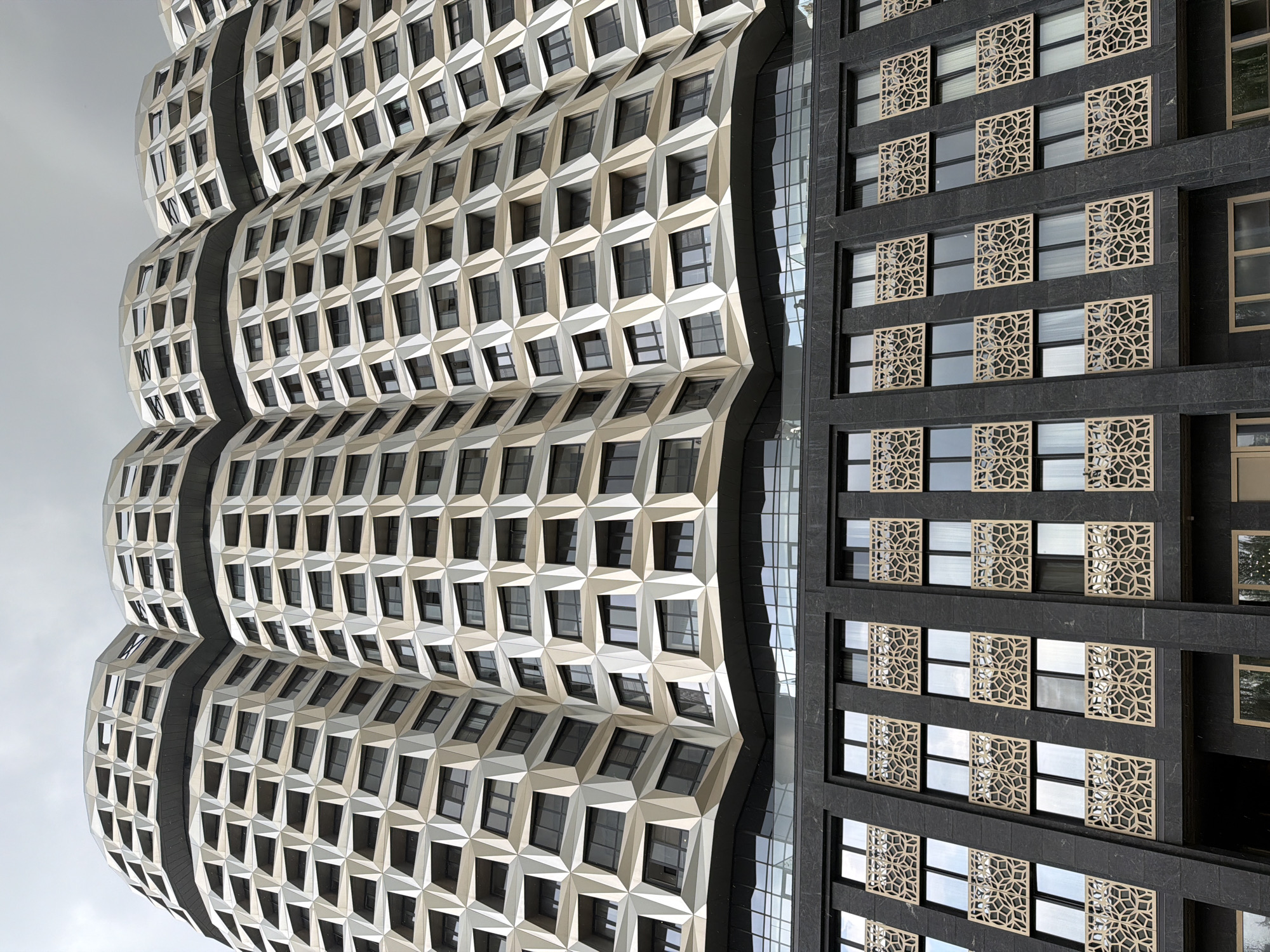 This is a low-angle, full-shot photograph of a modern architectural building in Moscow, Russia, under a cloudy sky. The building has a unique, undulating facade with a pattern of geometric shapes, appearing to be made of light-colored metallic panels, which are segmented by darker lines. Numerous windows are visible within these geometric sections. The upper portion of the building has a curved, almost organic shape, resembling stacked, rounded elements. Below this, a darker section of the building features rows of windows, each adorned with decorative, light-colored metal screens with intricate geometric patterns. The lower floors appear to be clad in a dark, textured material, possibly stone. The overall impression is one of contemporary and somewhat abstract design.