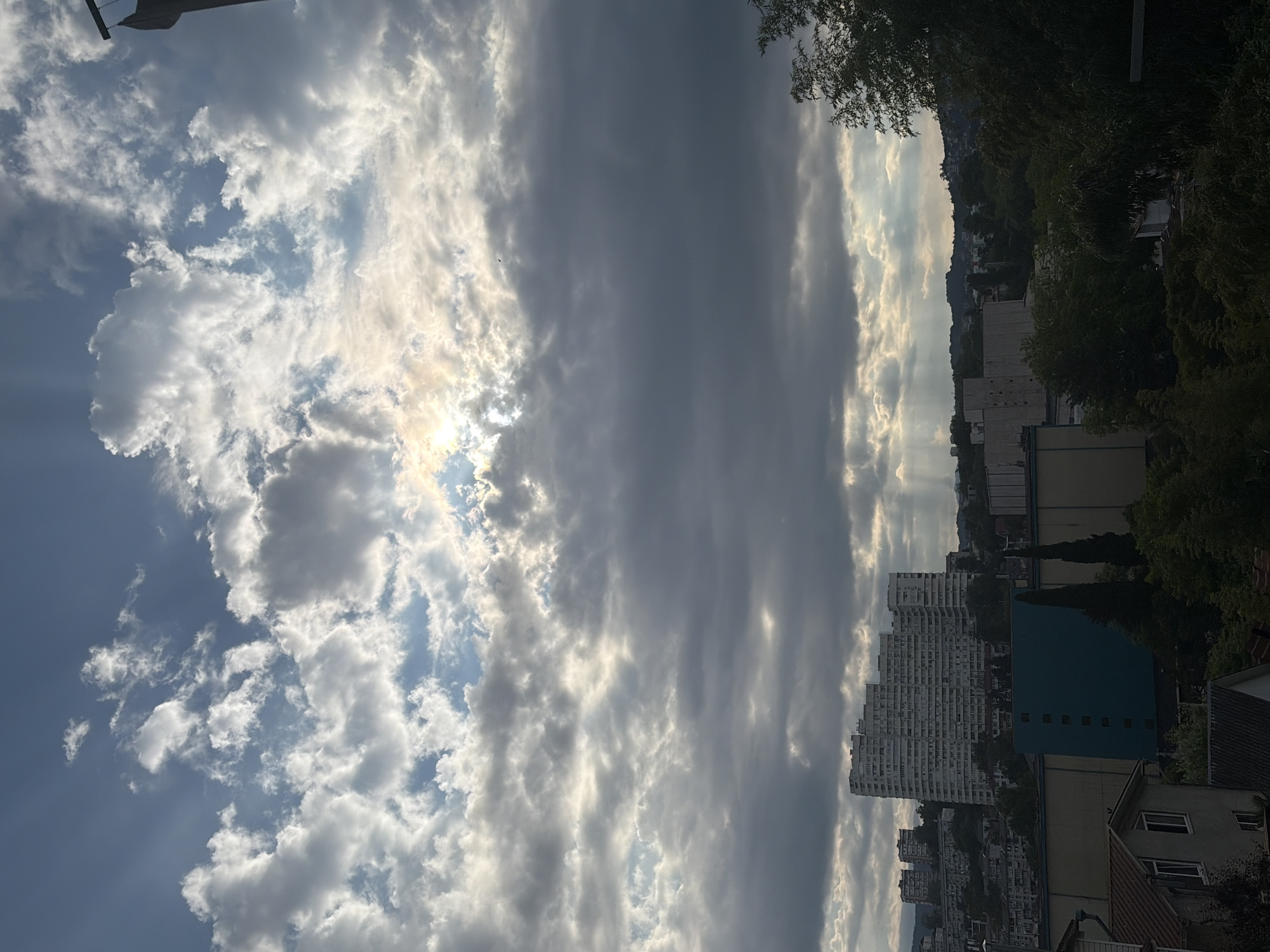 The image depicts a panoramic view of the city of Sochi, Russia, under a dramatic sky filled with broken clouds. Sunlight streams through gaps in the clouds, creating visible crepuscular rays that illuminate the scene. In the mid-ground, a large, curved, white residential building dominates the skyline, typical of Soviet-era architecture found in Sochi. Behind it, smaller buildings and a densely wooded hillside suggest the city's hilly terrain. The foreground reveals the tops of trees and parts of residential buildings, hinting at a higher vantage point from which the photo was taken. The overall impression is one of a partly cloudy day over a coastal city.