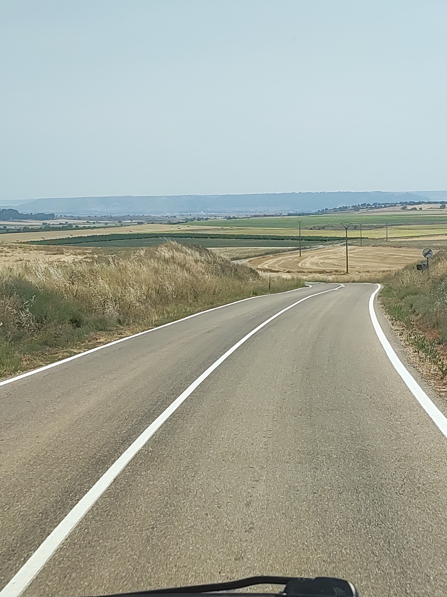 This is a photograph of a rural road winding through a landscape of agricultural fields under a clear sky. The road, paved with asphalt and marked with white lines, curves gently from the foreground into the distance. On either side of the road, dry grass and sparse vegetation line the shoulders. Beyond the immediate roadside, the landscape opens up to reveal fields with varied textures and colors, suggesting different crops or stages of harvest. Some fields appear golden brown, possibly harvested wheat or barley, while others are a darker green. In the middle distance, several utility poles with crossbars stand scattered across the fields, indicating the presence of electrical infrastructure. Further in the background, a hazy horizon is formed by rolling hills, also dotted with what appear to be trees or scattered vegetation. The overall impression is one of a dry, open countryside typical of a rural Spanish environment. The specific location is the city of Traspinedo, Spain.