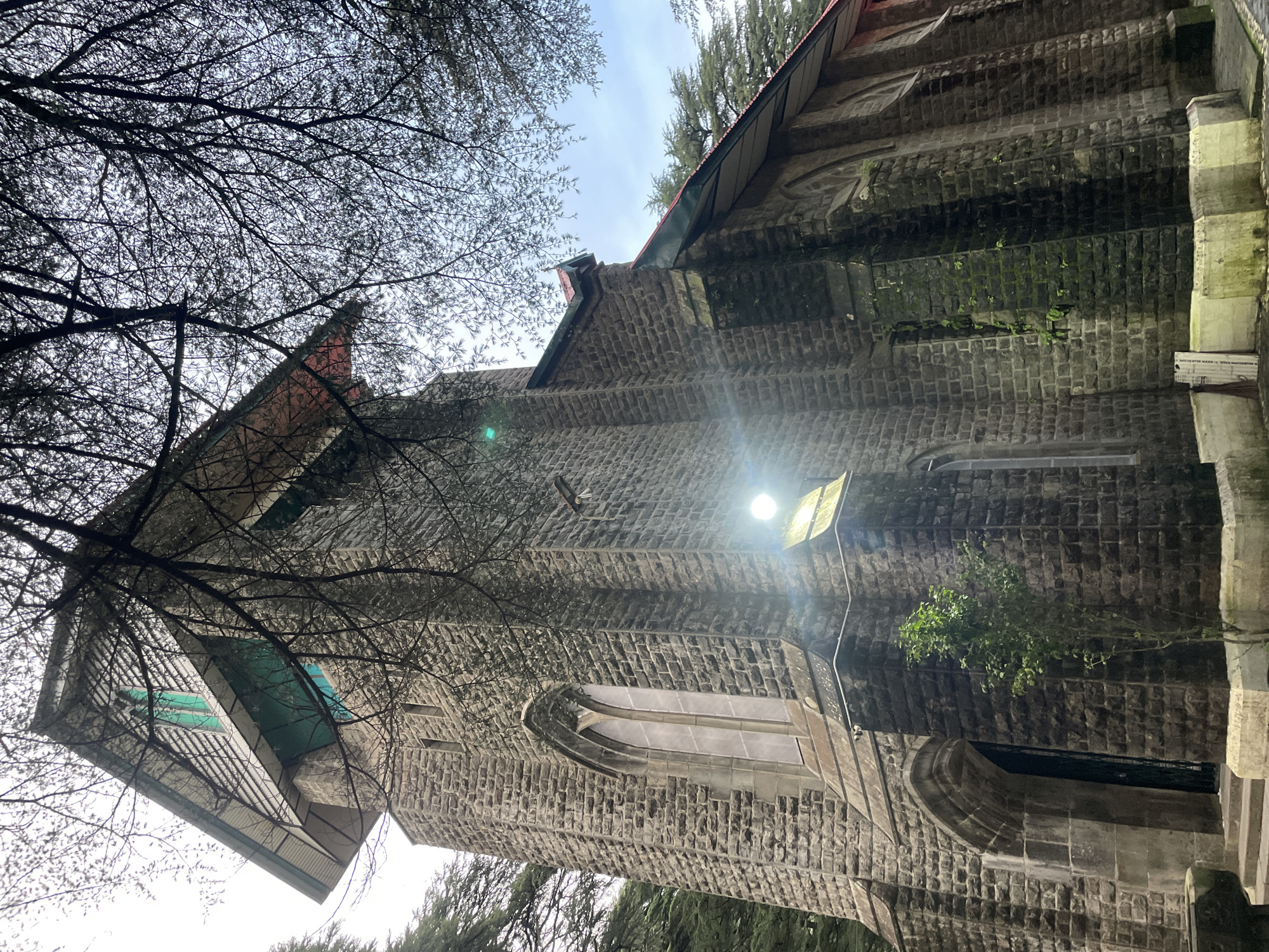 A low-angle shot captures a stone church tower in Dharamkot, India, viewed from below through the bare branches of a tree. The tower is constructed of rough-hewn grey stone blocks, with arched windows and a steep, gabled roof. The top of the tower features a lighter, almost white material with horizontal slats and two distinctive teal-colored windows. A single, thin tree grows near the base of the tower. A bright light is visible on the side of the tower, creating a lens flare. The sky above is overcast and pale. The image conveys a sense of history and perhaps solitude.