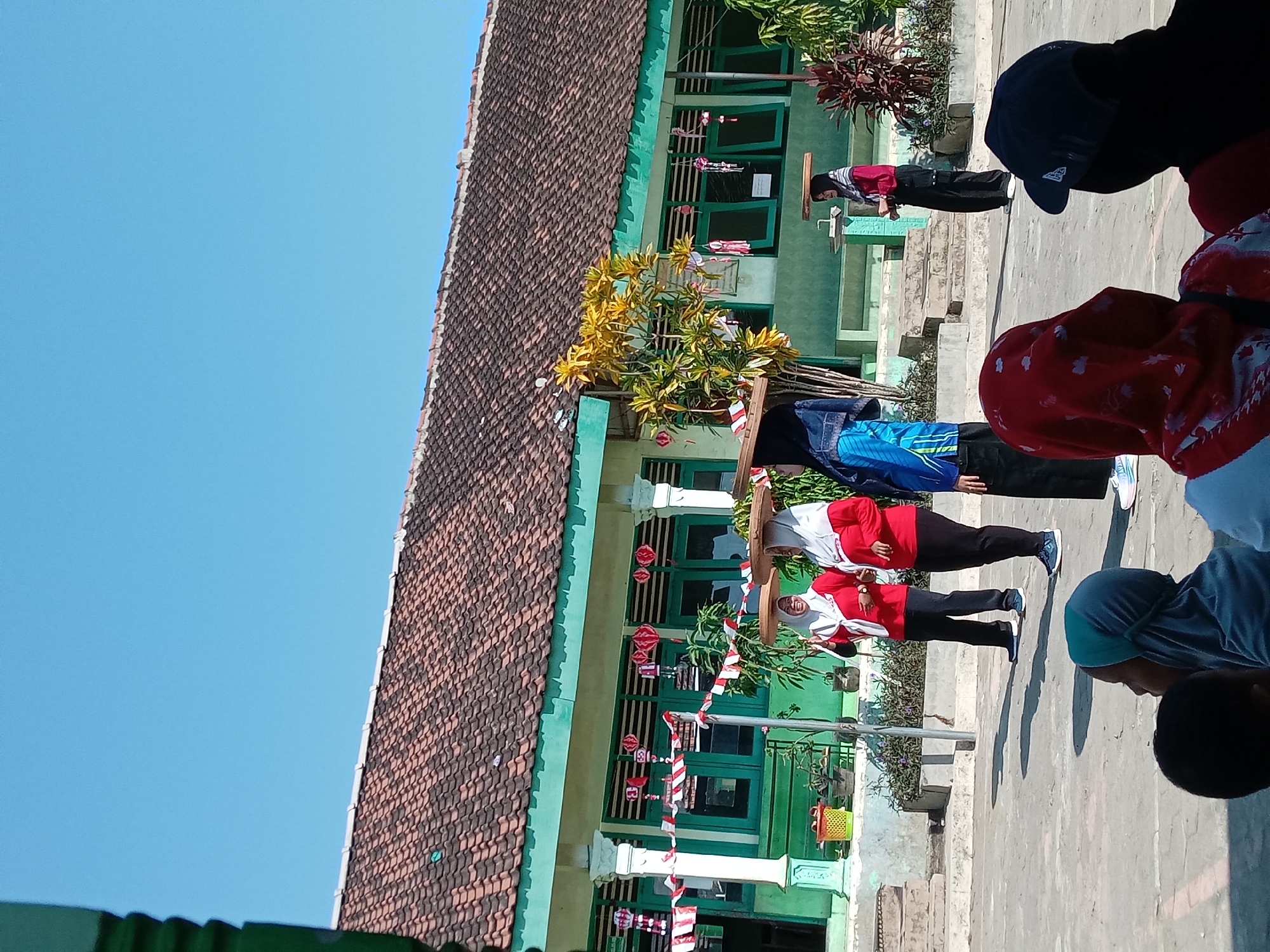In a courtyard in front of a school building in Ngompak, Indonesia, four young women in traditional Islamic headscarves and modest clothing are participating in a game or performance. They are carrying flat, round wooden trays on their heads, balancing them as they stand and move. Two women in red shirts and black pants stand closer to the foreground, with one smiling and gesturing. Further back, a woman in a blue and black patterned top and black pants, and another woman in a red top and black pants, also balance trays on their heads. The school building behind them is painted a light green and has a terracotta tiled roof. Red and white striped banners and paper lanterns are hung across the front of the building, suggesting a celebratory occasion. Several children and adults are seated in the foreground, watching the performance. The ground is paved with concrete, and a few trees and plants are visible. The sky is clear and blue.