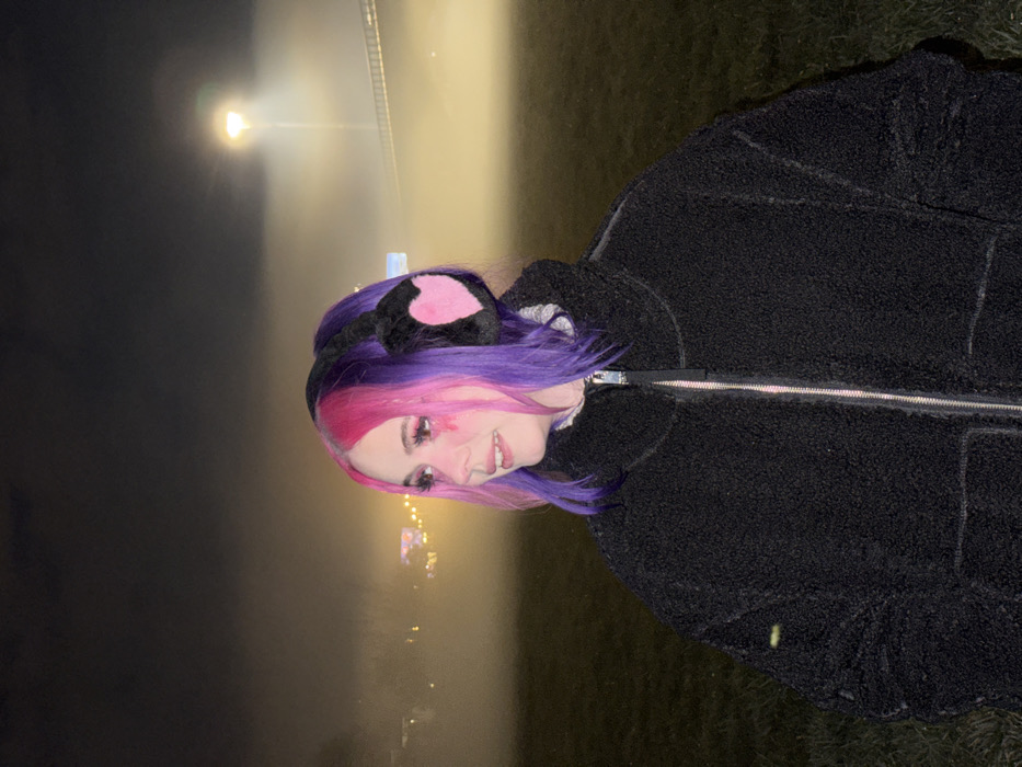 The image displays a single individual, appearing female, positioned centrally in a nighttime outdoor environment characterized by dense fog. The individual's hair is dyed in two distinct colors: bright pink on the left side and purple on the right. She wears black, plush ear muffs with pink heart-shaped centers and a black, textured, zippered jacket. Her makeup includes extended eyelashes and pink streaks descending from the outer corners of her eyes, and a small metallic labret piercing is visible on her upper lip. Her head is turned slightly to the left, exhibiting a subtle smile. The foreground consists of a dimly lit grassy field. The background is heavily obscured by fog, through which a tall stadium-style floodlight pole with a bright yellowish-white light is visible on the right, along with indistinct structural elements of a large facility. This scene is consistent with the grounds surrounding Maksimir Stadium in the City of Zagreb, Croatia.