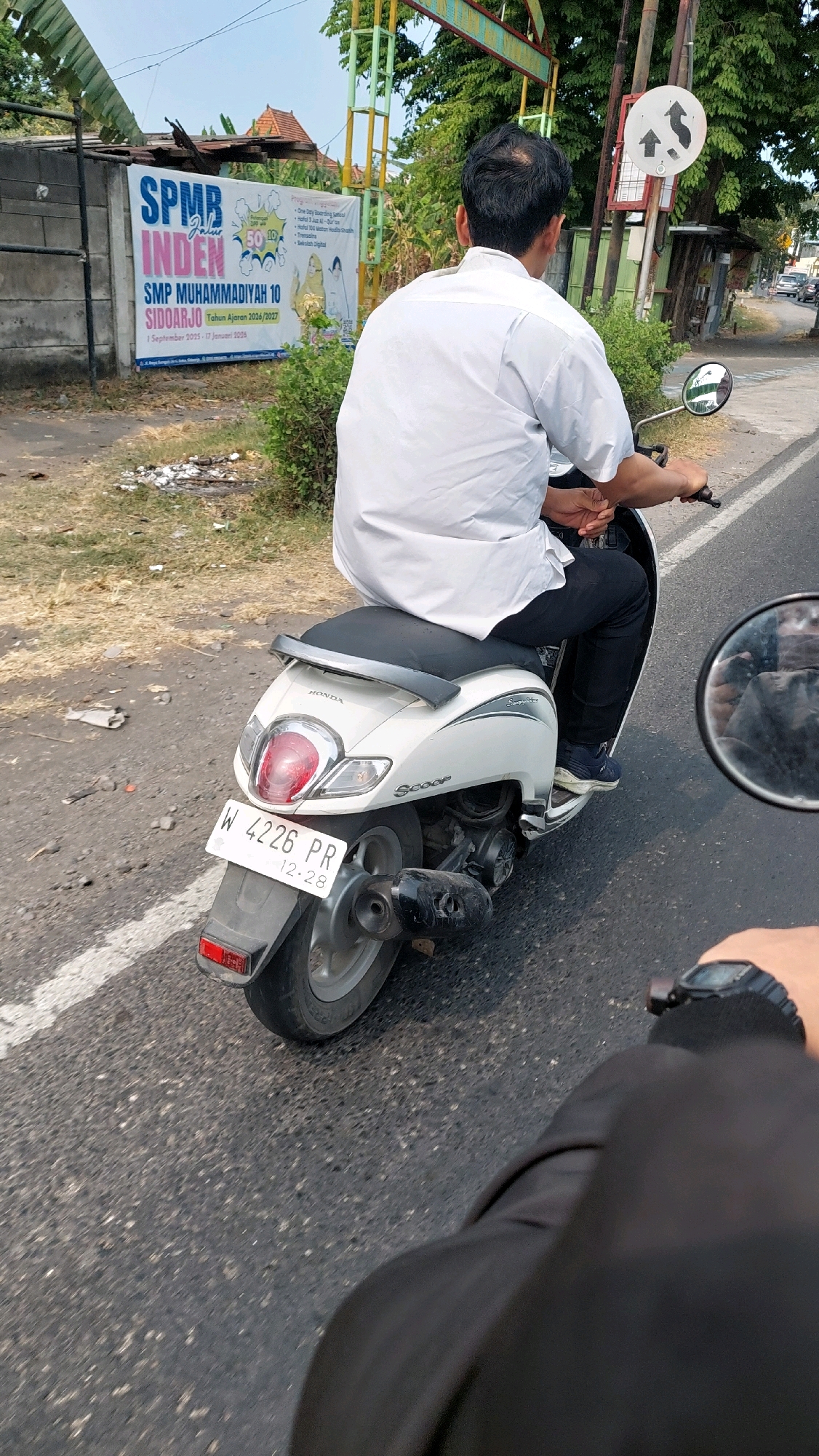 In Sidoarjo, Indonesia, a man wearing a white short-sleeved shirt and black pants rides a white Honda Scoopy motorcycle. He is seen from behind, facing away from the camera. The motorcycle has the license plate "W 4226 PR" and the registration sticker shows "12.28". He is on a paved road with a white line marking the lane. To his left, there is a banner that reads "SPMB INDEN SMP MUHAMMADIYAH 10 SIDOARJO". The background shows vegetation, a wall, and a street sign with directional arrows. The lighting suggests it is daytime. Another person's arm wearing a black jacket and a watch is visible in the foreground, indicating the photo was taken from another motorcycle.