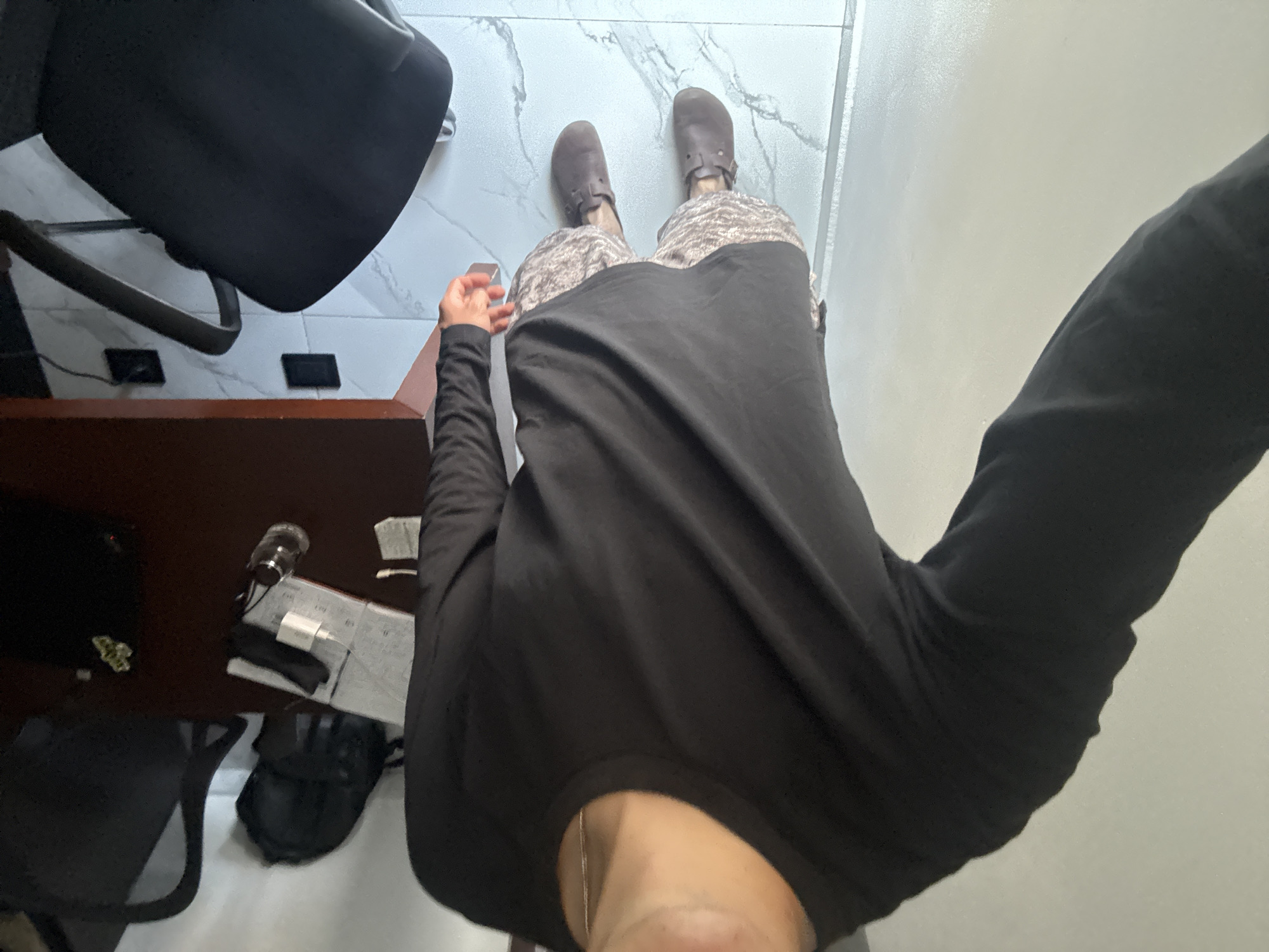 A first-person perspective image shows a person standing in what appears to be an indoor setting with tile flooring. The individual is wearing brown shoes, patterned pants or shorts, and a black long-sleeved shirt. In the background, there's a desk with items such as a lamp, papers, a phone charger, and a chair. The location appears to be a residential or office space in San Bernardino Tlaxcalancingo, Mexico.
