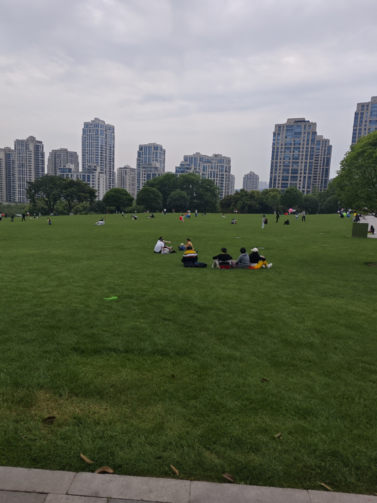 The image shows a large grassy field in Liangjiang, China, dotted with people engaged in various activities. Several groups are sitting on the grass, some appear to be picnicking. Individuals can be seen walking or standing in the distance. In the background are modern high-rise buildings, indicating a urban park setting. The sky is overcast. The ground appears to be well-maintained grass. A paved area is visible in the foreground.