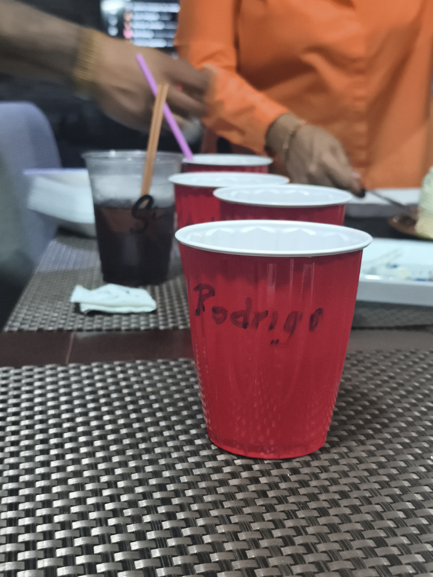 The image depicts a close-up view of a table setting, possibly at a gathering or event. There are three cups in the foreground. The foremost cup is red with the name "Rodrigo" written on it in black marker. Behind it are two other red cups, partially obscured. Further in the background, there is a plastic cup with a dark liquid and two straws. The table is covered with a woven placemat with a grid pattern. In the background, there are people, one wearing an orange shirt and another partly visible, suggesting a social setting. The location is not identifiable from the image.