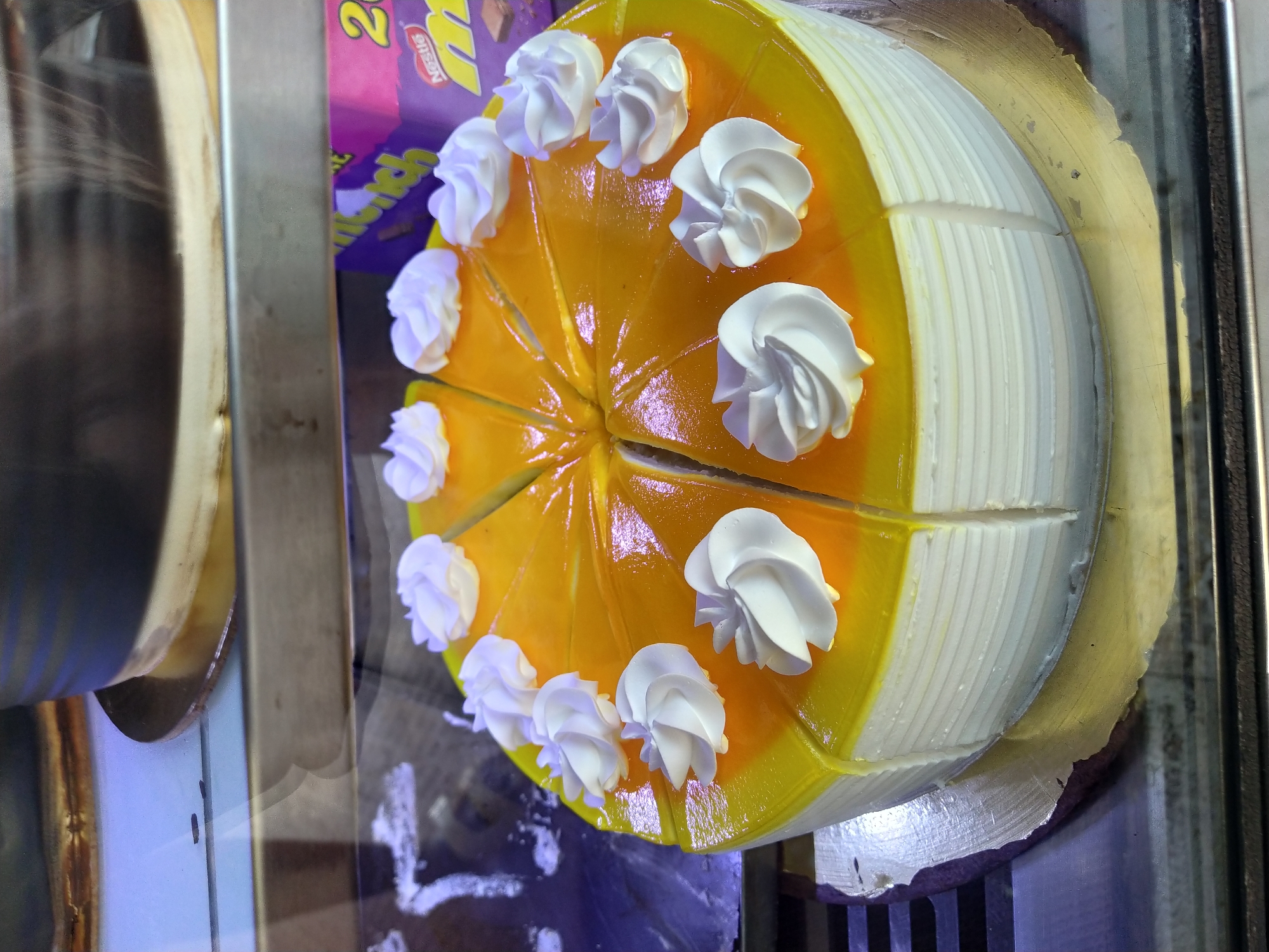 The image shows a sliced cake with yellow jelly topping and white cream rosettes, likely in a display case. The cake is cut into several slices and appears to be ready for sale. In the background, there's another cake and part of a purple wrapper, possibly candy or another food item. The picture was taken in Mapusa, India.