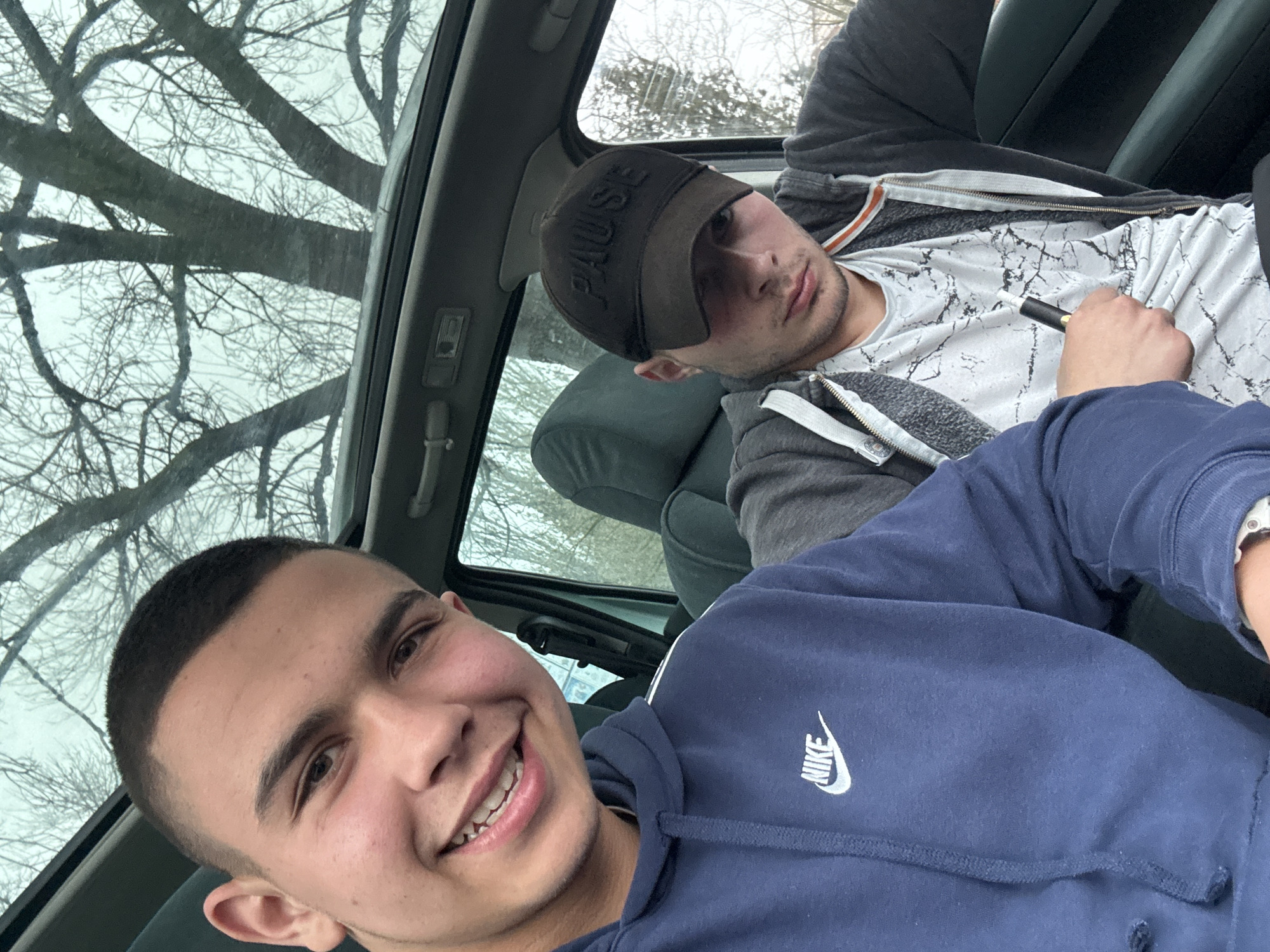 Two young men are sitting in a car. The man in the foreground is wearing a blue Nike hoodie and smiling. The man in the background is wearing a black hat with the word "Pause" on it, a white t-shirt, and a grey zip-up hoodie. He is holding a black pen or vape pen. The car has a sunroof, and bare tree branches are visible through it. The location is unknown, so I cannot include Topoli, Bulgaria.