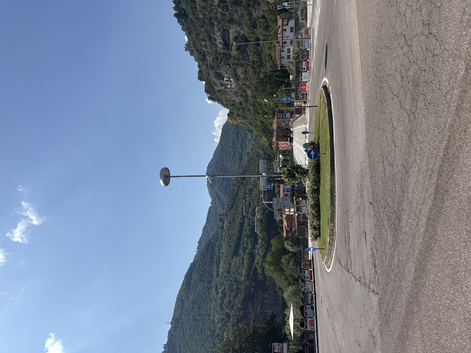 The image captures a wide-angle view of a roundabout in the city of Trento, Italy, set against a backdrop of lush green mountains. The sky is a clear, bright blue with a few wispy clouds. In the foreground, the asphalt of the roundabout shows signs of wear with visible cracks. A modern streetlamp stands tall in the center of the roundabout, illuminating a well-maintained green island with a small cluster of trees and bushes.
Several signs are visible around the roundabout. Traffic signs with blue arrows indicate the direction of travel. Further into the roundabout, on the right side, a pedestrian crossing is marked. On the left side of the roundabout, a "BAR" sign is prominently displayed above outdoor seating, suggesting a commercial establishment. Various advertisements, some featuring "CONAD" and "COOP SUPERSTORE," are positioned along a barrier.
In the mid-ground, vehicles are navigating the roundabout, and a few pedestrians can be seen walking. Buildings with red and orange facades are nestled among the trees on the slopes of the mountains. The mountain slopes themselves are densely covered with vegetation, with some rocky outcrops visible. The overall scene depicts a typical day in a town where urban infrastructure meets natural landscape.