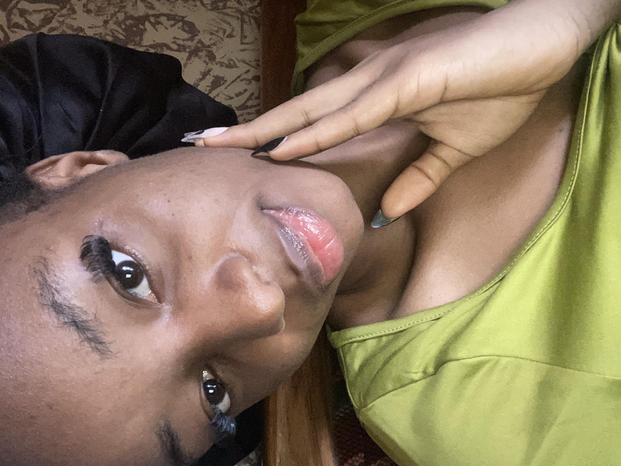 A close-up, high-angle selfie shows a young Black woman with dark skin, her face angled towards the camera. She is wearing a lime green top and a black satin bonnet. Her left eye is in focus, with prominent false eyelashes and a reflection of light visible in her pupil. Her lips are slightly parted, appearing glossy and pink. Her right hand is raised, with manicured nails featuring a black and white design, touching her cheek. The background is blurred, showing a patterned fabric and a wooden surface. The image appears to be taken indoors, possibly in a home environment in Jos, Nigeria, given the context provided by the prompt.