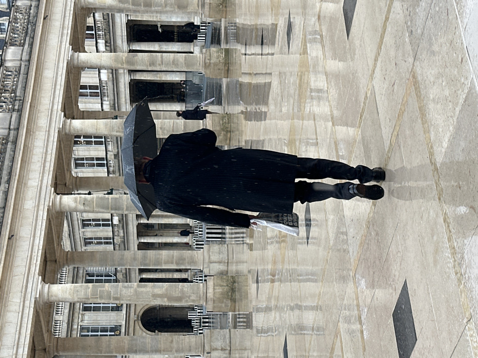 The scene depicts an adult male walking away from the camera in the Cour d'Honneur of the Palais Royal in Paris, France. It is daytime and raining, evidenced by the wet, highly reflective paved ground and visible rain streaks. The man wears a dark long coat, dark trousers, and dark shoes, holding an open black umbrella above his head with his right hand. In his left hand, he carries a white paper shopping bag with dark text. The background features the neoclassical stone architecture of the Palais Royal, characterized by numerous tall columns, arched entrances, and windows. Further in the mid-ground, the distinctive black and white striped art installation known as Les Deux Plateaux (Colonnes de Buren) is visible. Several other indistinct figures can be seen in the distance, some also under umbrellas.