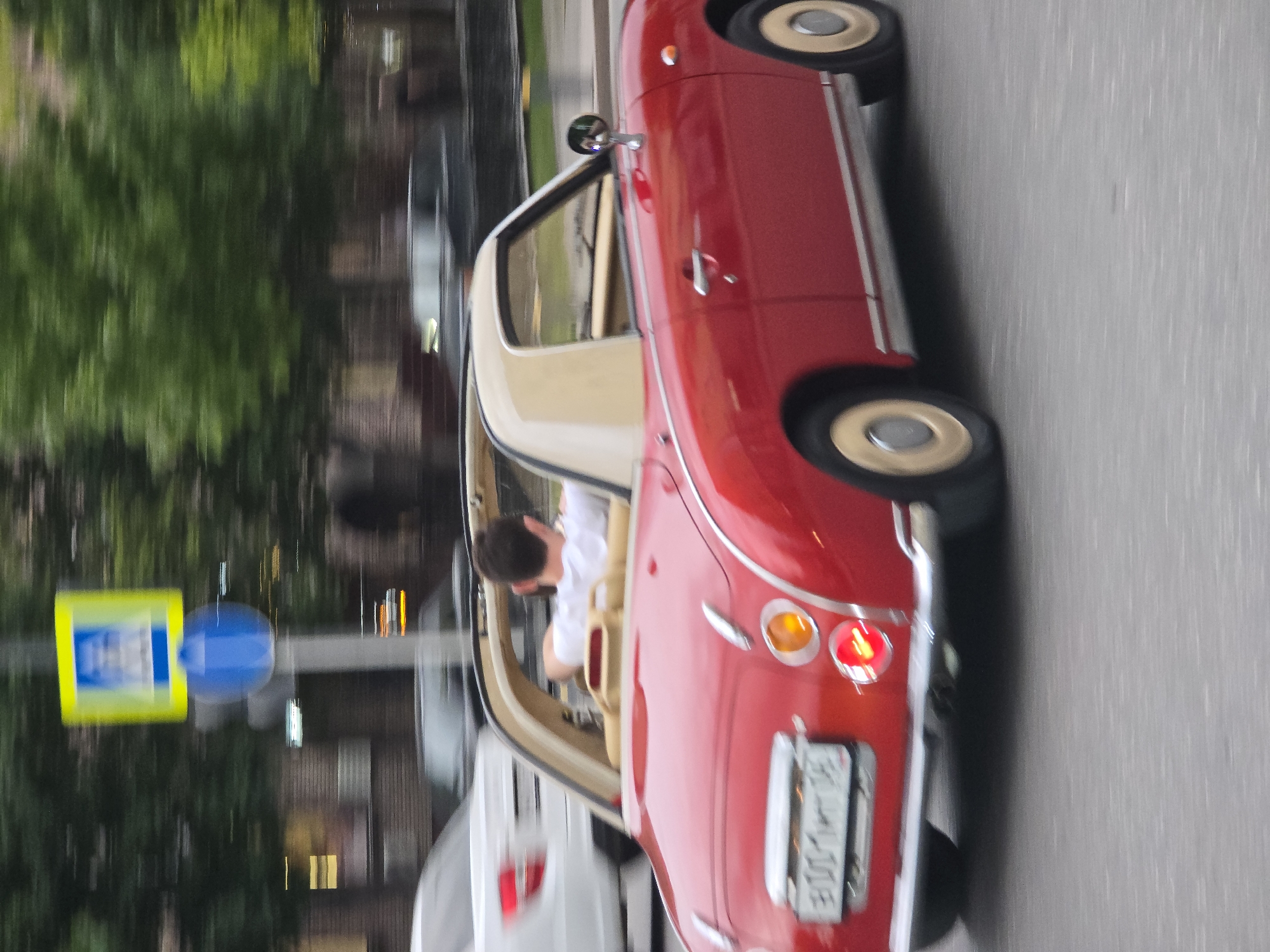 A blurry eye-level shot, taken outside in the city of Unknown, Unknown, shows a red Nissan Figaro driving past a crosswalk sign, captured with motion blur. The car is viewed from its right side, with a man visible behind the wheel. The car has a tan roof and cream-colored wheels with tan centers. The license plate is partially visible as "A001." A blurred white car follows behind. The background includes trees and city lights. The pavement of the road is gray.