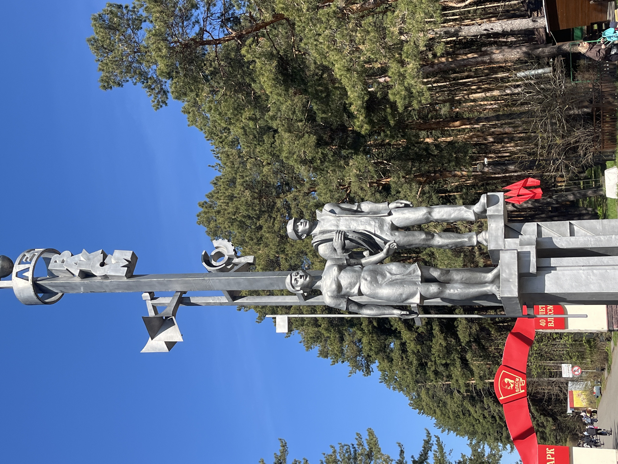 The image features a monument in Ulyanovsk, Russia, set against a backdrop of tall pine trees and a clear blue sky. The monument consists of two silver-colored statues of working-class individuals mounted on a gray metal structure. The structure also includes a ring with stylized lettering and additional abstract shapes. At the base of the monument is a red archway-like structure, adorned with lettering in Russian. Pedestrians and foliage are visible in the lower right corner, indicating the monument's location within a park or public space.