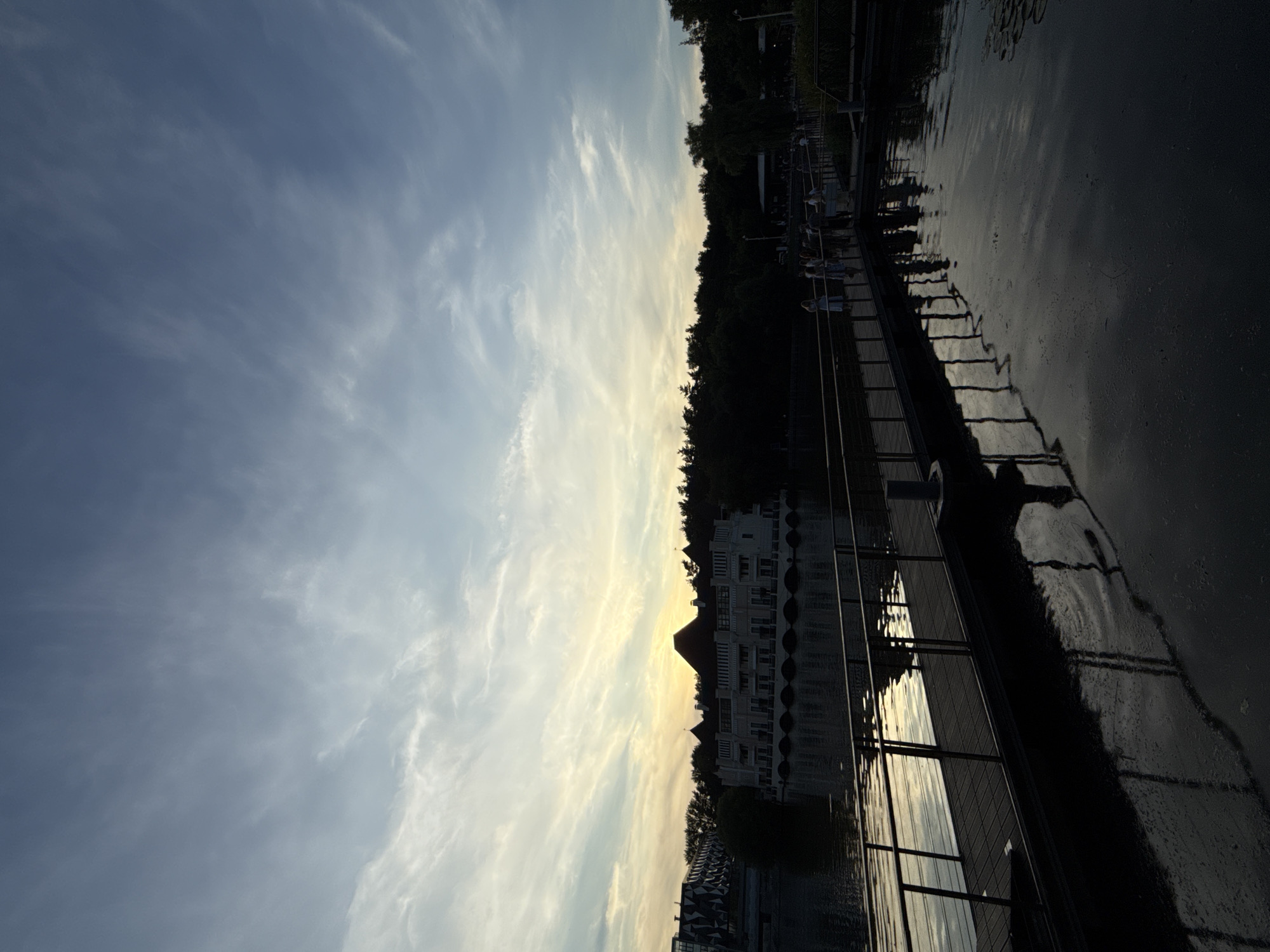 The image depicts a pier extending over a body of water, likely a lake or river, in Moscow, Russia. The scene is set during sunset, with the sky displaying a gradient of blues and yellows, illuminated by wispy clouds. On the opposite bank of the water, a light-colored building with arched windows and a dark, peaked roof is visible, surrounded by lush trees. To the left of this building, a more modern structure with a geometric facade can be seen. Several individuals are present on the pier, appearing to be strolling or conversing. The water reflects the sky and the structures, creating a serene and tranquil atmosphere. The pier is equipped with a metal railing and its surface appears to be made of wood or composite material. The overall impression is one of a picturesque urban park or waterfront area.