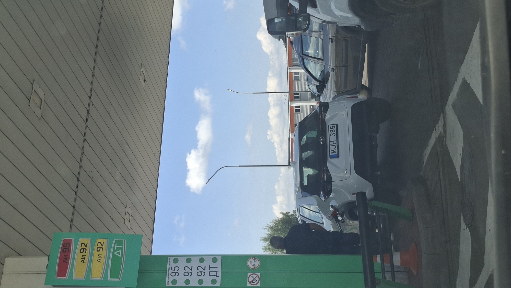 The image shows a gas station in Myadzyel, Belarus. A white Toyota Yaris is parked at a gas pump, with a fuel nozzle inserted into the car's fuel tank. A man in a dark patterned shirt stands next to the car, observing the refueling process. The gas station features a green pylon with prices for different fuel grades displayed, including AI-95, AI-92, and DT. The sky is blue with scattered white clouds. In the background, other cars and buildings are visible, suggesting a typical urban or semi-urban environment.