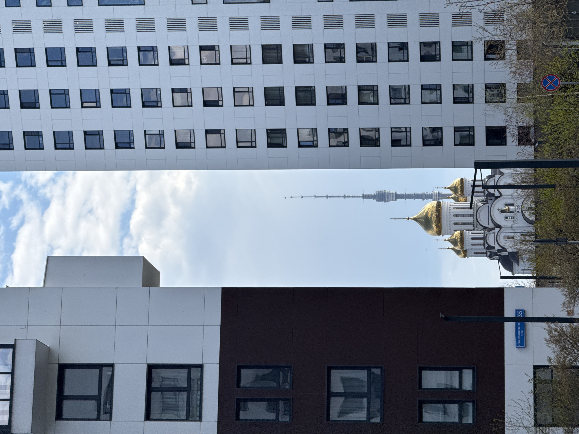 The image shows a view in Moscow, Russia, featuring buildings and landmarks. The composition is somewhat unconventional, with the scene potentially rotated for artistic effect.
At the top of the frame is a modern, white office building with a grid of square windows. In the mid-ground is the skyline, showcasing the golden domes and spires of a Russian Orthodox church, along with the distinctive Ostankino Tower in the distance. The foreground contains another modern building with a combination of white and dark brown facade sections and square windows. A street sign indicating "Сенгилеевская Улица 35" (Sengileevskaya Street 35) is visible, helping pinpoint the location. A "No Stopping" road sign and trees with budding leaves suggest an urban environment during the transition from winter to spring. There are no people visible in the image.