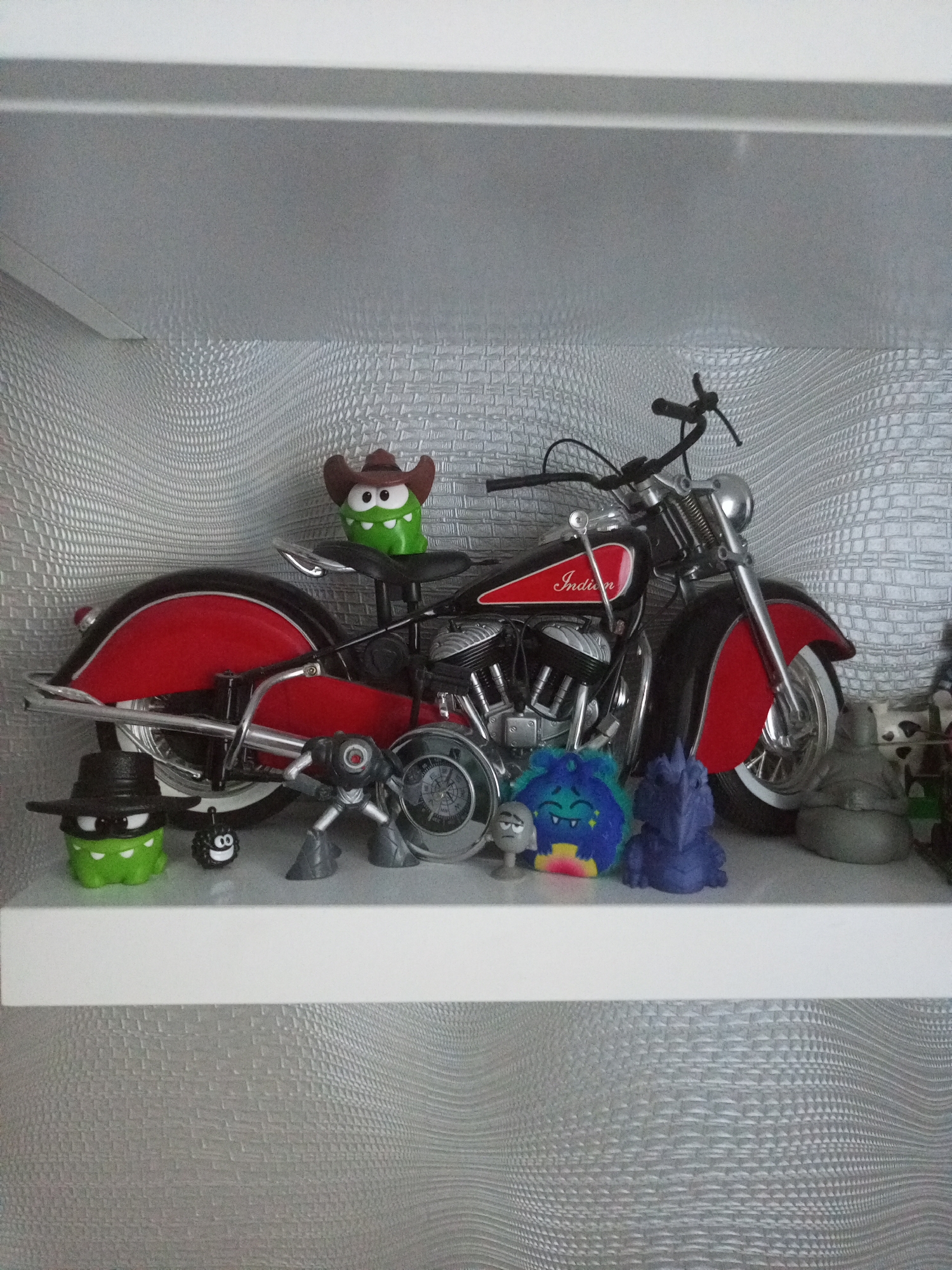 In Homyel, Belarus, an indoor shelf displays various decorative items. A red and black Indian motorcycle model is the central object, with a green character wearing a cowboy hat perched on its seat. To the left of the motorcycle, a green character in a black hat and a small black character with a smiley face are positioned. To the right of the motorcycle, there are several small figurines: a gray robotic figure, a blue fuzzy monster, a gray dragon-like figure, and a gray seated figure. The shelf is illuminated, and the background features a textured, light-colored wall.