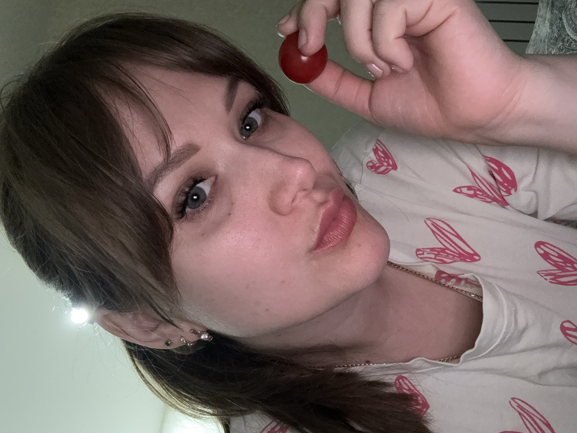 A woman is holding a small, round, red object (likely a cherry tomato) between her thumb and index finger. She has shoulder-length brown hair with bangs and is wearing multiple earrings in her left ear. She's wearing a light-colored shirt with pink wing designs and a gold necklace. The background is indistinct, and there is no discernible information to determine if the location is Saratov, Russia.
