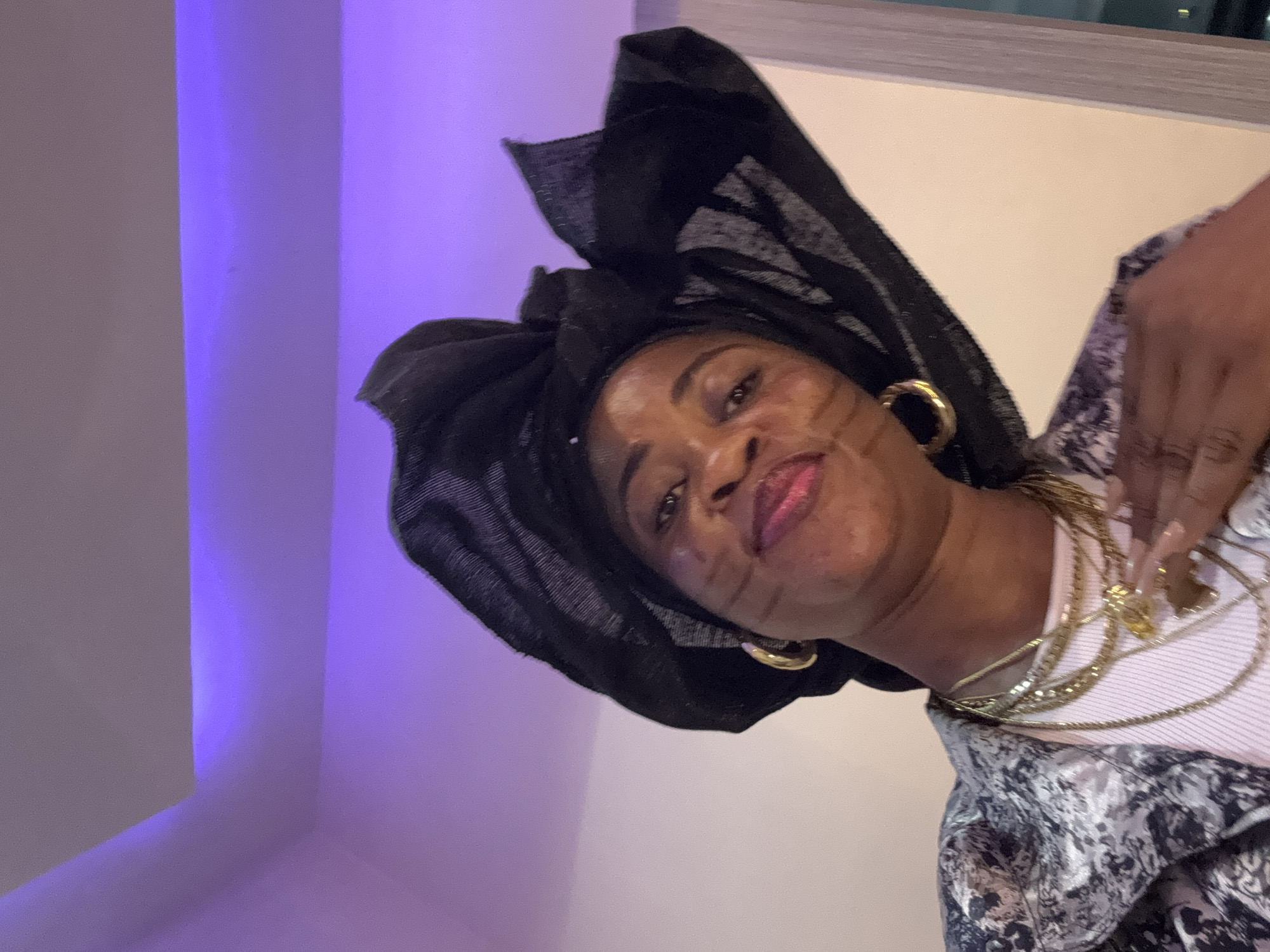 A close-up, head-and-shoulders shot shows a Black woman in Ajara, Nigeria, wearing a black headscarf, gold hoop earrings, a patterned jacket over a pink top, and several gold necklaces. She is looking directly at the camera with a slight smile and is touching her neck with her right hand. The background features a purple-lit ceiling and a beige wall with a window.