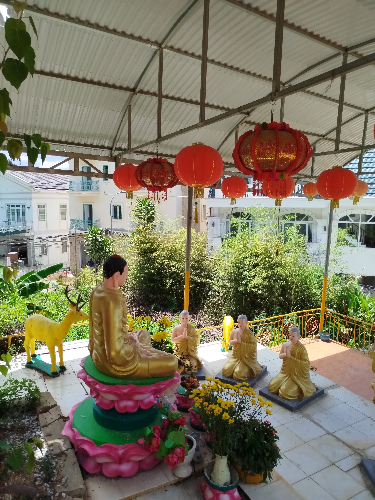The image shows a courtyard or covered patio area in Da Lat, Vietnam. Underneath a metal frame roof with hanging red lanterns, there's a Buddhist shrine or display. A golden statue of Buddha sits on a lotus flower base. To the left of the Buddha is a golden deer statue. Three smaller golden statues of monks kneeling in prayer are positioned in front of the Buddha. Flowers are placed in front of the statues. The background features greenery, trees, and a multi-story building.