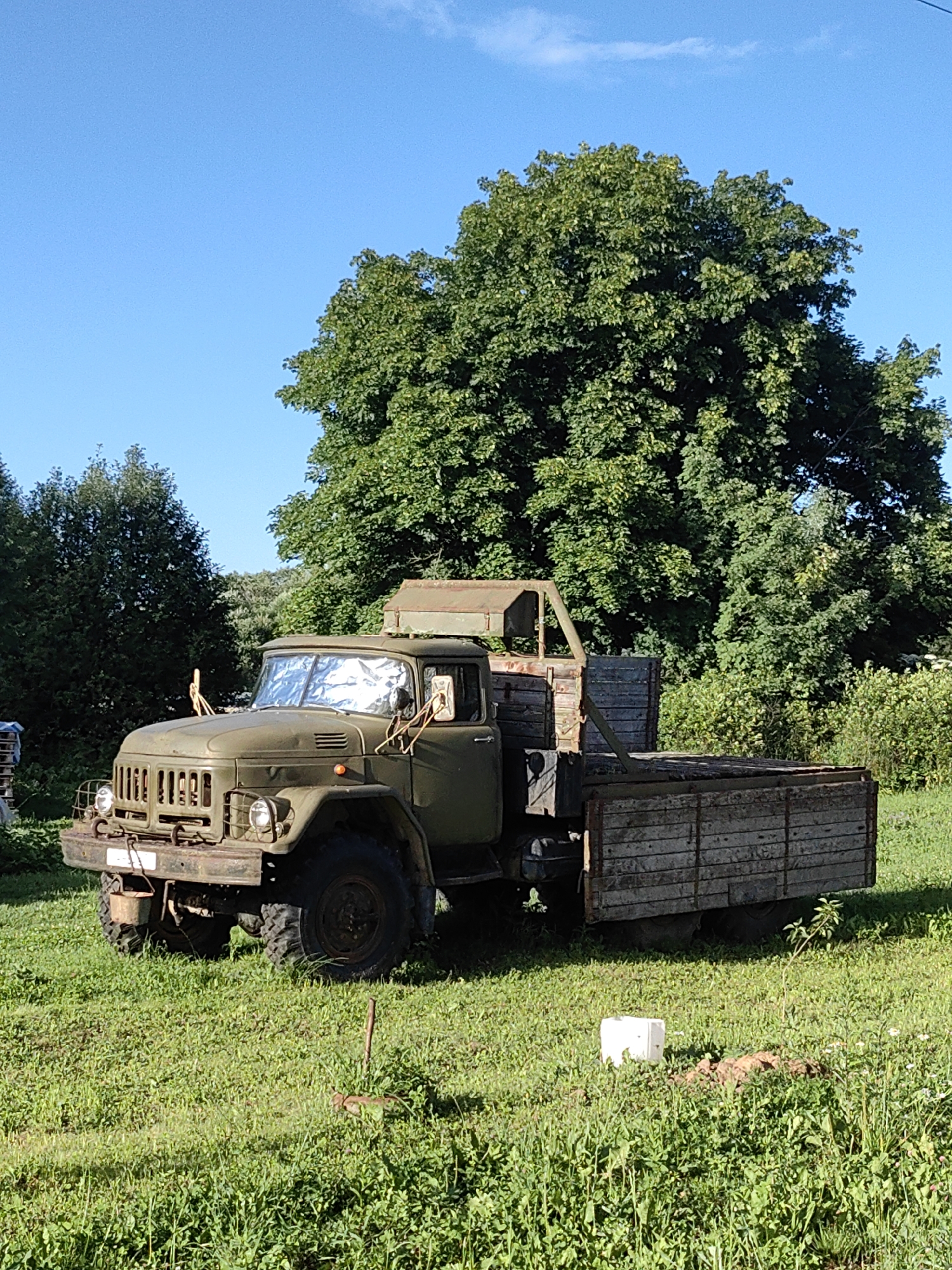 A vintage, olive-green truck sits parked on a grassy field under a clear blue sky with some scattered clouds. The truck, which appears to be a utility vehicle with a wooden bed, has a reflective sunshade covering its windshield. The background features lush green trees and foliage, suggesting a rural or park-like setting. To the right of the truck, a small mound of dirt and a white object are visible in the grass. The overall scene is one of stillness and apparent disuse of the vehicle.