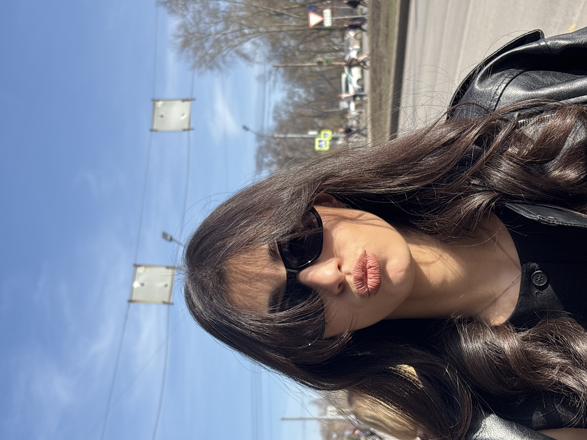 A woman with long brown hair and sunglasses poses for a selfie on a street in Perm, Russia. She wears a black leather jacket. In the background, street signs, trees, and buildings are visible under a clear blue sky.