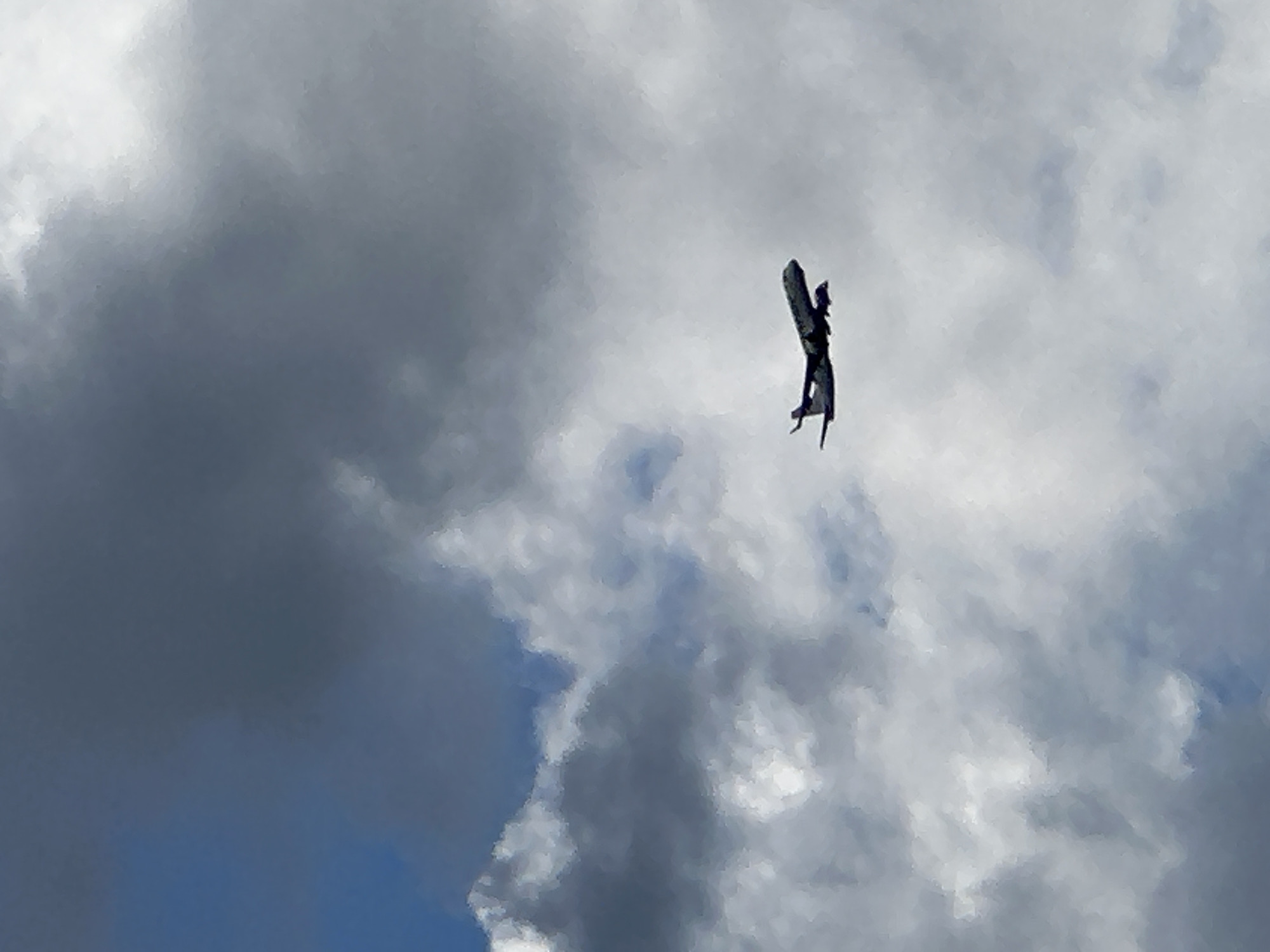 A silhouetted airplane is shown in flight against a backdrop of dark and light clouds. The airplane is positioned in the lower right quadrant of the image, with its nose pointing slightly upwards and to the right. The fuselage and wings of the aircraft are visible. The sky is filled with a mixture of dense, dark clouds and lighter, more diffused clouds, creating a dramatic and somewhat turbulent atmosphere. The lighting suggests it could be overcast or nearing sunset. The image was captured in an unknown location.