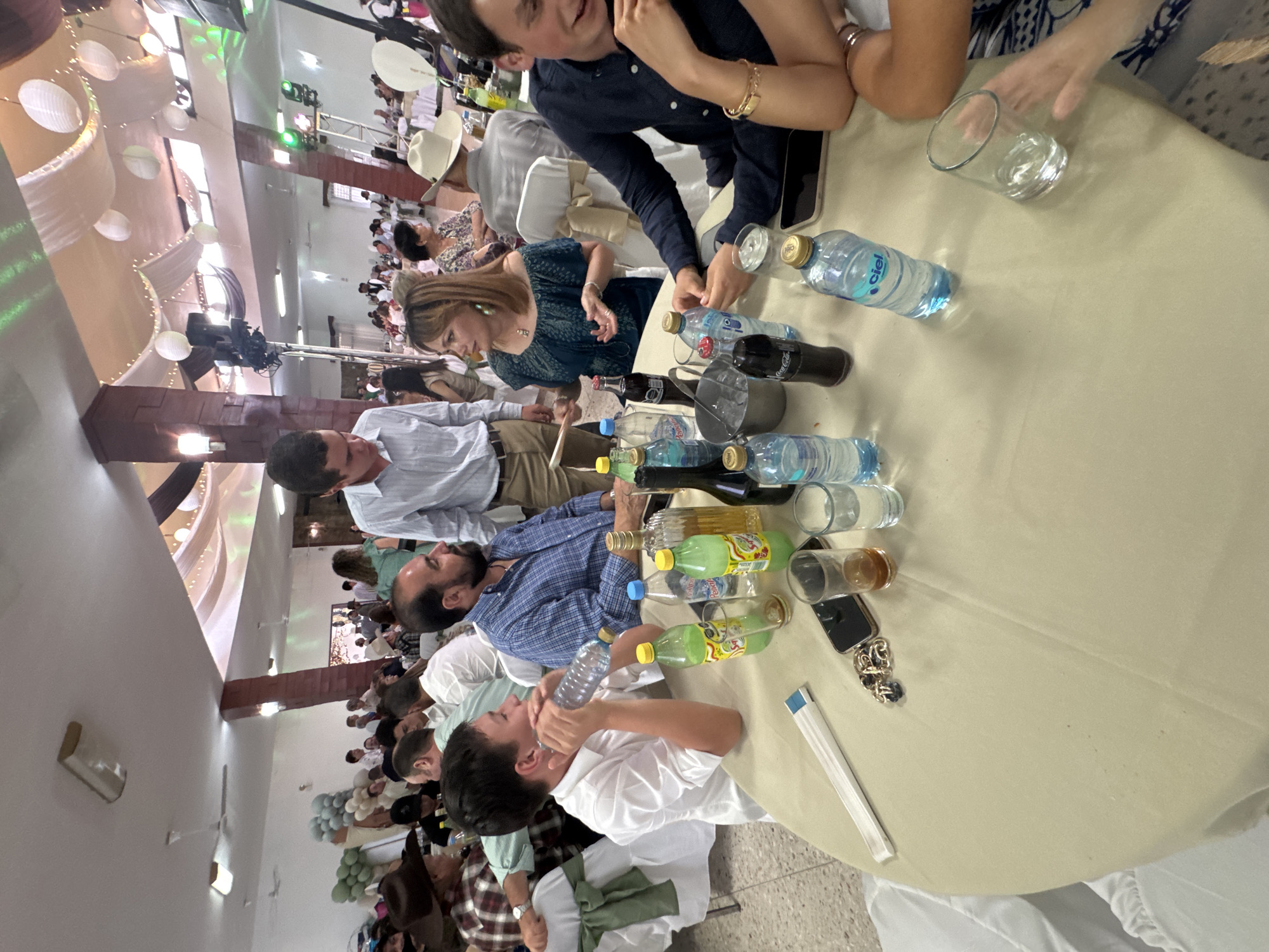 A slightly high-angle, medium indoor shot depicts a group of people seated around a table at an event or gathering. The location is identified as Chipilo de Francisco Javier Mina (Chipilo), Mexico. The people are diverse, with varying ages and appearances. The individuals at the table are dressed in smart casual attire.
The table is covered with a light-colored tablecloth. It is laden with bottles of water and beverages. There are also glasses and personal items like phones and jewelry scattered across the table.
In the background, more people are seated at other tables in a large room. Decorations such as white drapes, lights, and balloons adorn the space. The overall impression is that of a celebratory occasion.