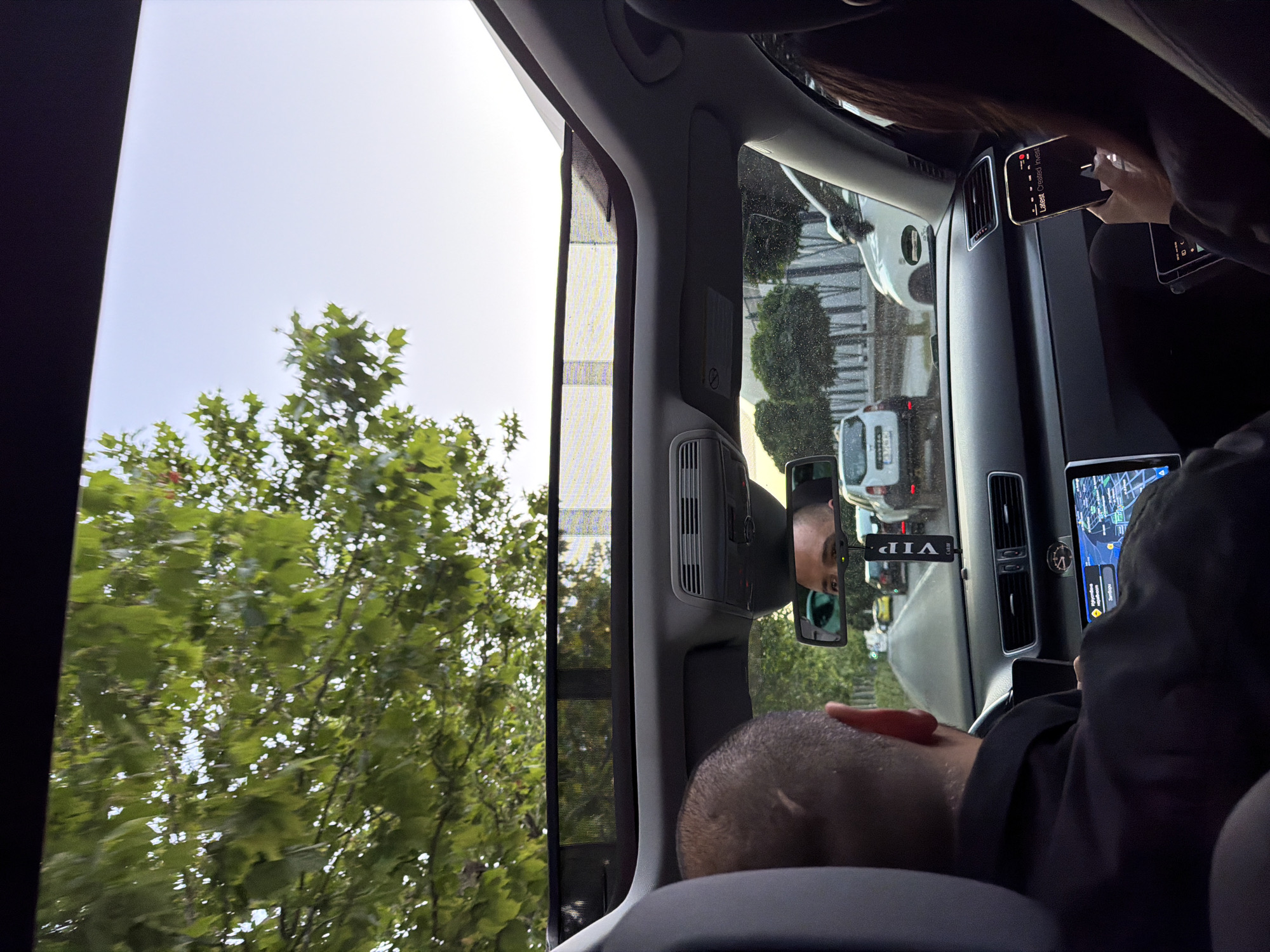 The image depicts the interior of a car with a sunroof open, revealing green foliage and a pale sky. The viewpoint is from the back seat, looking forward. The head of a man with a shaved head is visible in the foreground, with his hands on the steering wheel. A digital navigation screen is mounted on the dashboard. In the rearview mirror, the face of a man is reflected. A black "VIP" air freshener hangs from the mirror. The car is in traffic, with other vehicles visible on the road ahead, including a white SUV. Trees line the street. The scene appears to be in Burgas, Bulgaria, as indicated by the license plate of a white vehicle visible in the background which reads "Н 7146 BC". The overall impression is a snapshot of a journey within the city.