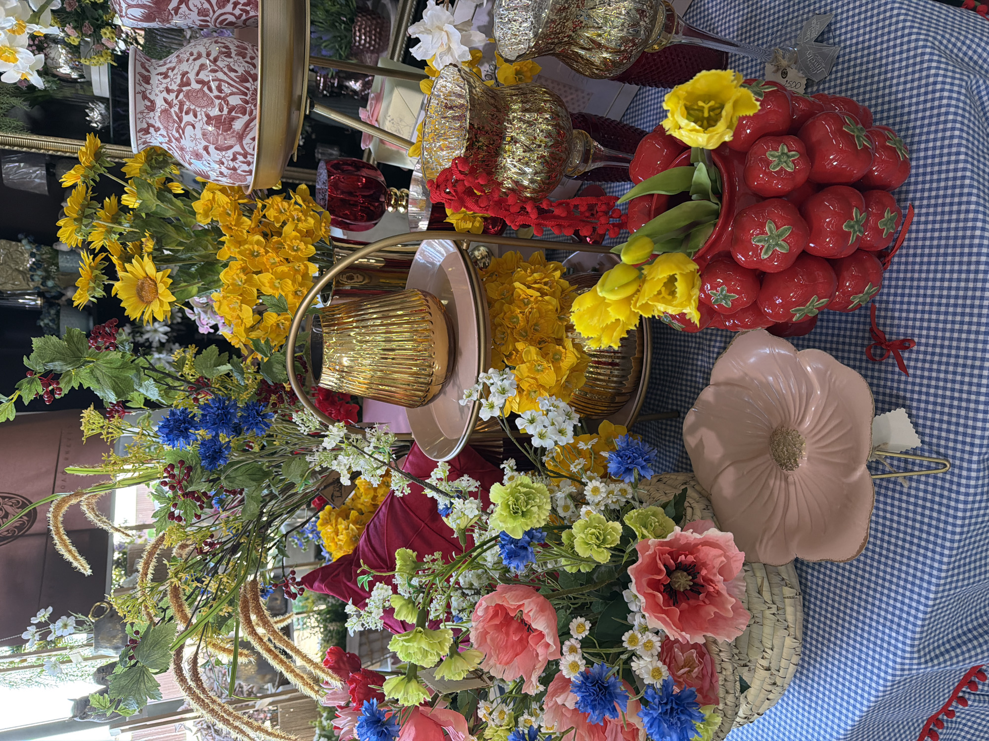 The image displays an indoor retail presentation of artificial flowers and decorative items on a table in Votkinsk, Russia. The table is draped with a blue and white checkered tablecloth, accented with a red pom-pom trim along its front edge.
In the foreground, to the left, a woven basket contains an arrangement of artificial flowers, notably large pink poppies, blue cornflowers, small white daisies, and lime green filler blooms. Adjacent to this, a large, matte light pink ceramic dish, shaped like a poppy flower, rests on a golden metal stand. Centrally, a red ceramic vase, textured with numerous individual ceramic strawberries, holds several artificial yellow tulips with green stems. A red ribbon is tied around the vase's base. Behind the strawberry vase stands a two-tiered golden metal display unit; its top tier holds a fluted golden metallic vase, and its bottom tier supports another similar vase surrounded by artificial yellow flowers. A deep red, possibly fabric, item is partially visible behind this display.
Further back, a dense, mixed arrangement of artificial foliage and flowers includes prominent brownish-gold grassy plumes, green leaves with red berries, blue cornflowers, and various yellow and white flowers. To the right, a tall, clear glass goblet-style vase with a textured golden finish features a red pom-pom garland draped over its side. Next to it is another textured golden glass vase. A white and red patterned ceramic vase, adorned with birds and floral motifs, rests on a golden tray. The background reveals additional retail merchandise, including dark red textured glass goblets, gift boxes, and mirrored surfaces within a dimly lit interior, suggesting a shop environment. No people are visible.
