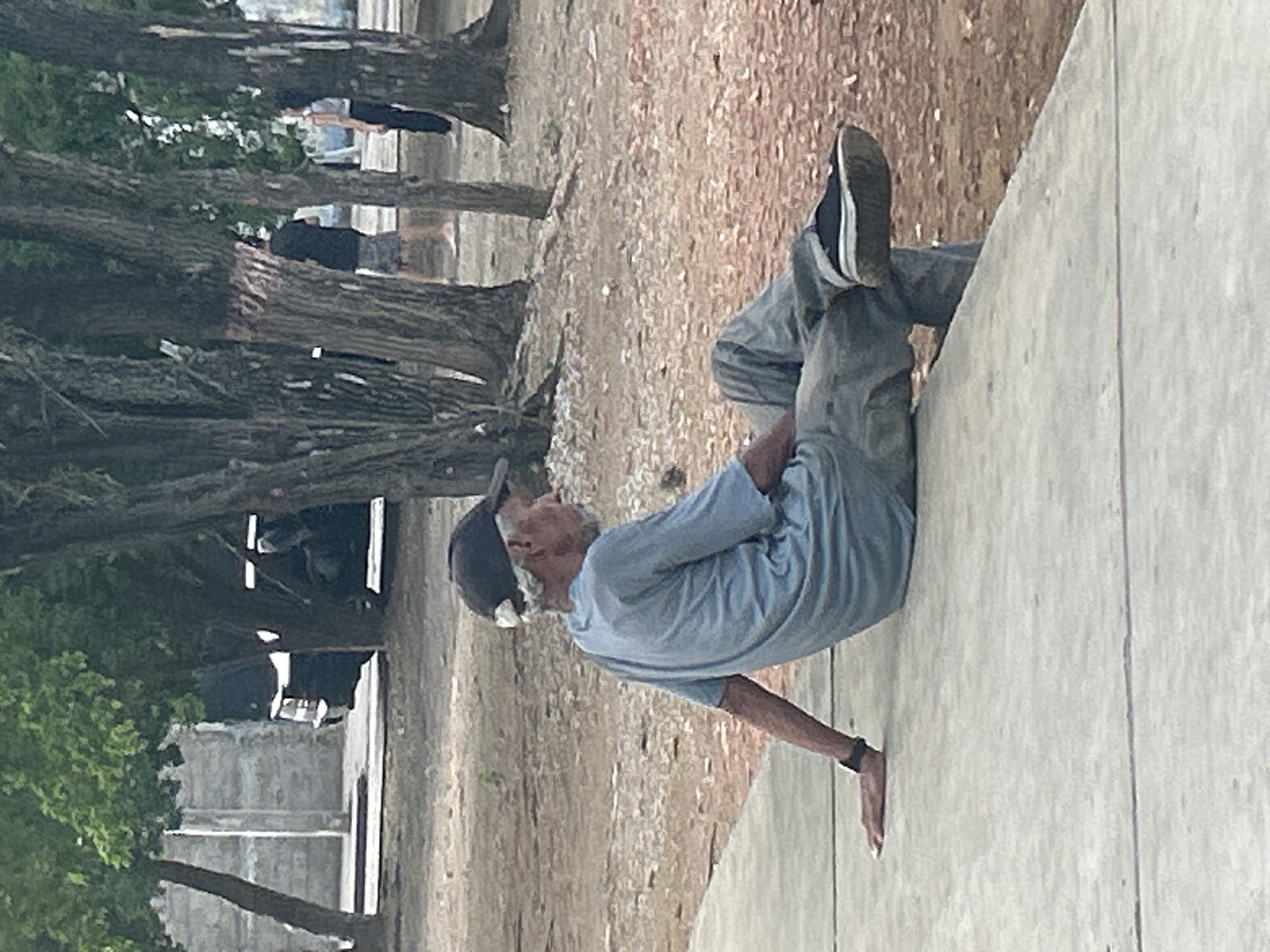The image depicts an elderly man with graying hair and a beard, wearing a blue t-shirt, grey pants, and a dark baseball cap, sitting on a concrete ledge outdoors. He is positioned with his legs crossed and his left arm resting on the ledge. The background suggests a park or outdoor public space with trees, dirt ground, and possibly some distant buildings or vehicles. The man is the primary subject, appearing to be resting or observing his surroundings. The overall scene is casual and unposed, conveying a sense of everyday life in an urban or semi-urban environment. The location is identified as Santa Ana, Venezuela.