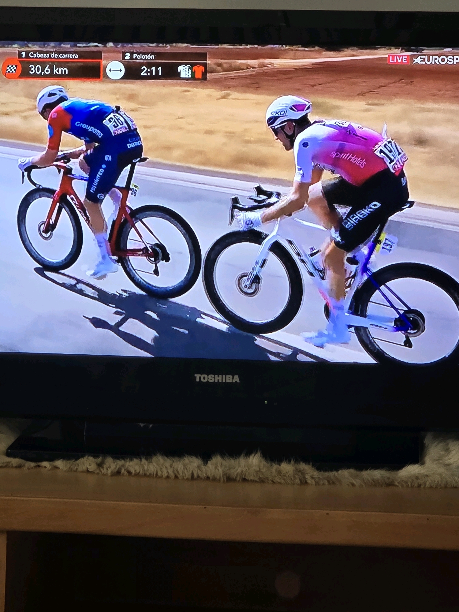 The image shows a close-up view of a television screen displaying a cycling race. Two cyclists are prominently featured, riding on a paved road. The cyclist on the left wears a blue and red racing suit and rides a red bicycle. The cyclist on the right wears a pink and white racing suit and rides a white bicycle. Both cyclists are wearing helmets and appear to be in motion.
At the top of the screen, a race information banner displays "1 Cabeza de carrera 30,6 km" on the left, indicating the leader and distance remaining. On the right, it shows "2 Pelotón 2:11" with icons representing a jersey and a rider. The "LIVE EUROSPORT" logo is visible in the upper right corner.
The background of the race scene shows a reddish-brown, dusty terrain, consistent with a desert or arid environment. The foreground includes a black television bezel with the "TOSHIBA" logo visible. Below the television, a wooden surface is visible with a fluffy, light-colored material draped over it. The overall lighting suggests an outdoor event. There is no discernible information in the image to indicate the location being the city of El Mourouj, Tunisia.