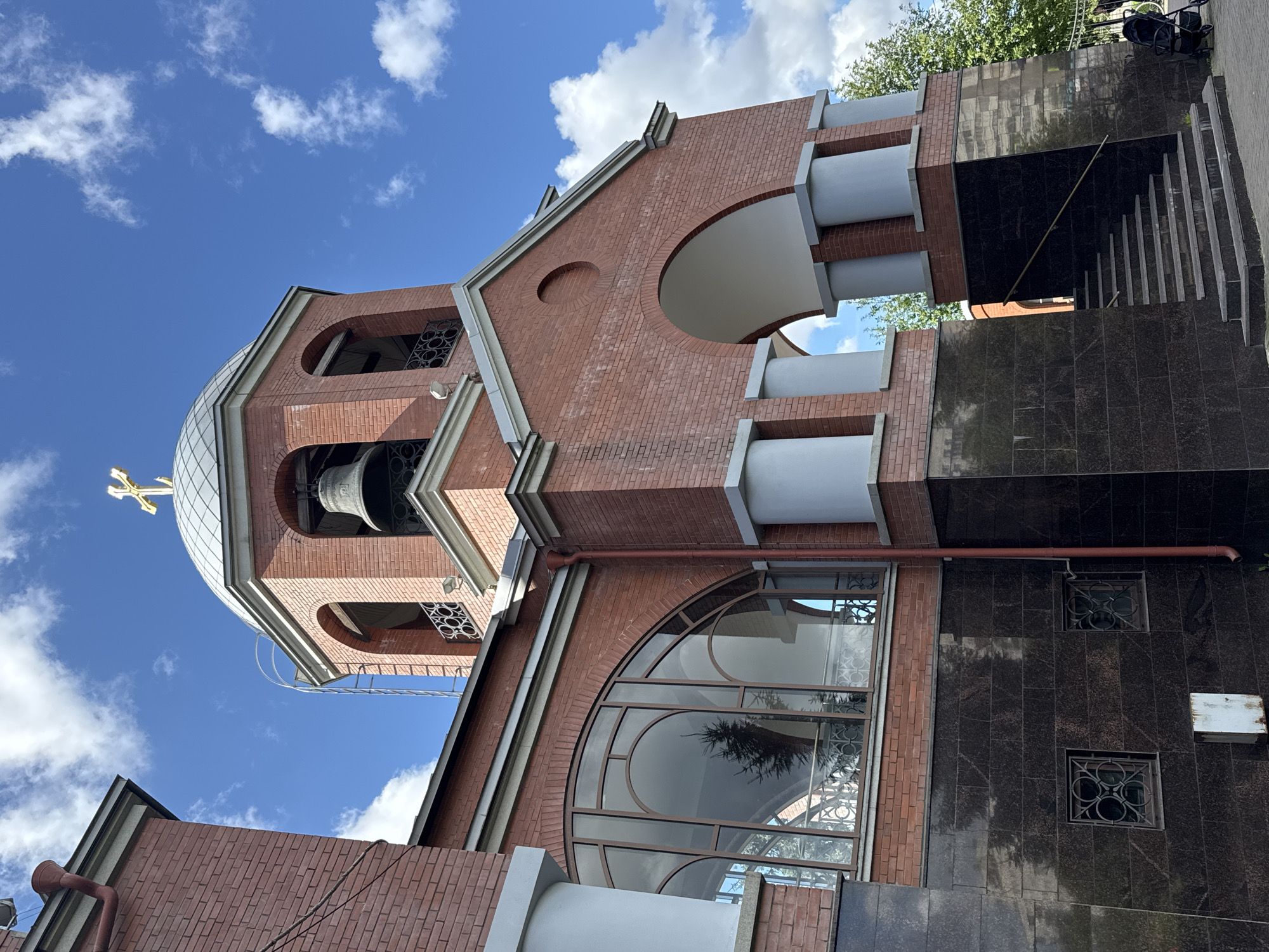 This image shows the exterior of the Church of the Resurrection of Christ, located in Saint Petersburg, Russia. The church is constructed of red brick and features a prominent bell tower topped with a golden cross. The bell tower has arched openings where a large bell is visible, and decorative metalwork. Adjacent to the bell tower is a large arched entryway with white pillars. A distinctive arched window with multiple panes is visible on the side of the building. The lower portion of the church is clad in dark granite. Steps lead up to the arched entryway. The sky is blue with scattered white clouds, and some greenery is visible in the background. A stroller is parked near the steps on the right side of the image.